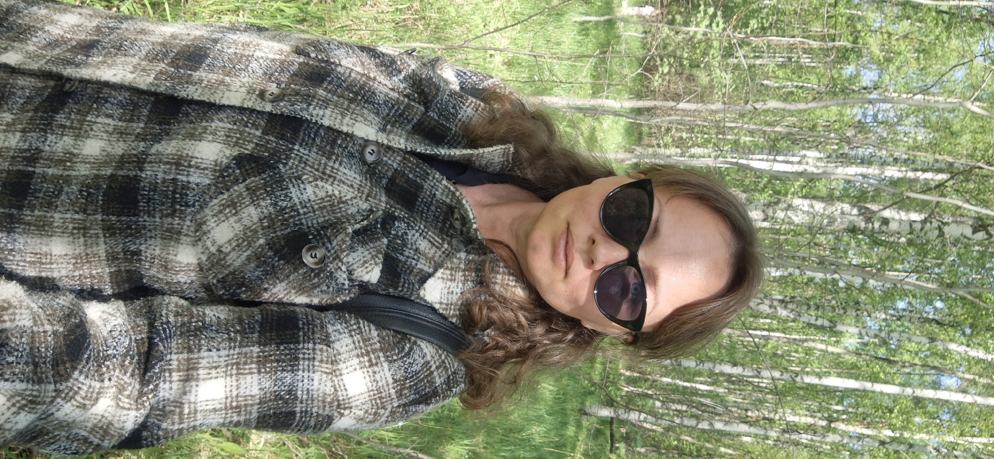 A woman with long, light brown hair and sunglasses is lying on her back in a grassy area near a birch forest. She is wearing a brown and white plaid jacket. The photo was taken in Innolovo, Russia.