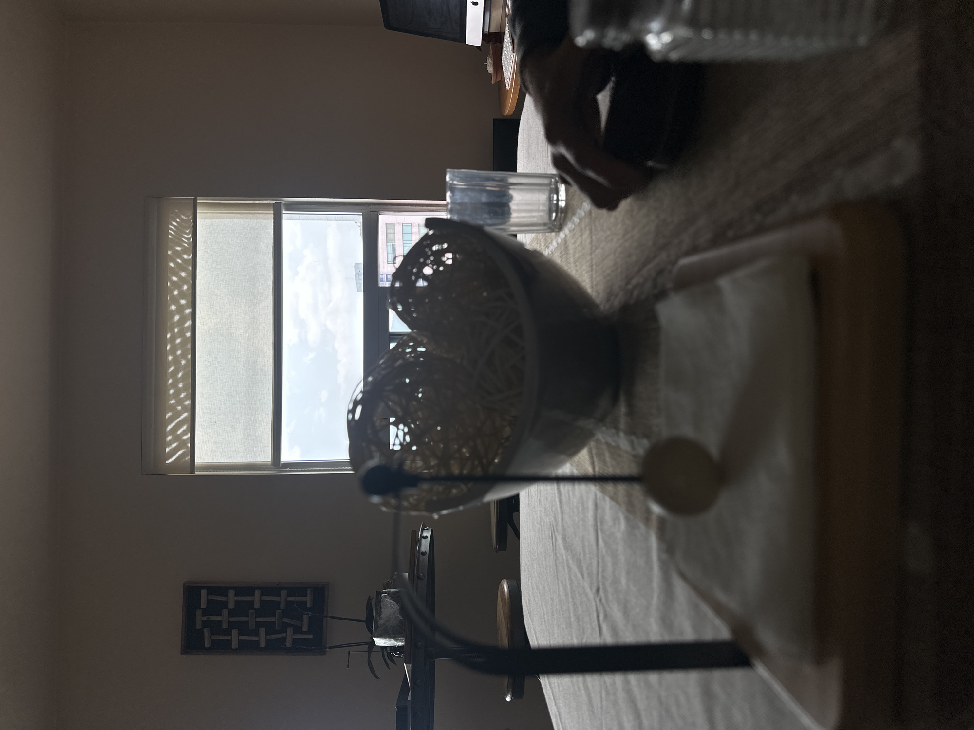 The image shows a dimly lit interior scene. A window with partially drawn blinds is visible on the left. A table is covered with a textured tablecloth and holds several items: a bowl, a drinking glass, and a stack of napkins in a napkin holder. A computer monitor is visible in the background. It is not possible to determine the location or any specific details related to Jesús del Monte, Mexico from the image alone. There are no people visible, and no actions or interactions are depicted.