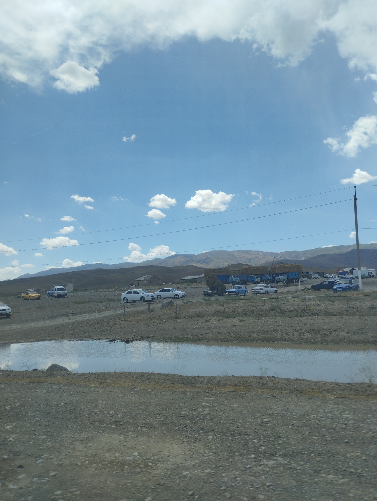The image depicts a rural landscape under a partly cloudy blue sky. In the foreground, there is a gravelly area with a shallow pool of water reflecting the sky. Beyond the water, a dirt road leads to a collection of buildings and vehicles. Several cars, including a yellow sedan, a white sedan, and a blue sedan, are parked in an open area. Further in the background, a line of large blue trucks loaded with hay bales is visible, suggesting agricultural activity. Mountains form the distant horizon, their slopes covered in dry vegetation. A utility pole with wires stretches across the scene, indicating the presence of infrastructure. The overall impression is of a functional, possibly agricultural, outpost in a remote, mountainous region of Uzbekistan.