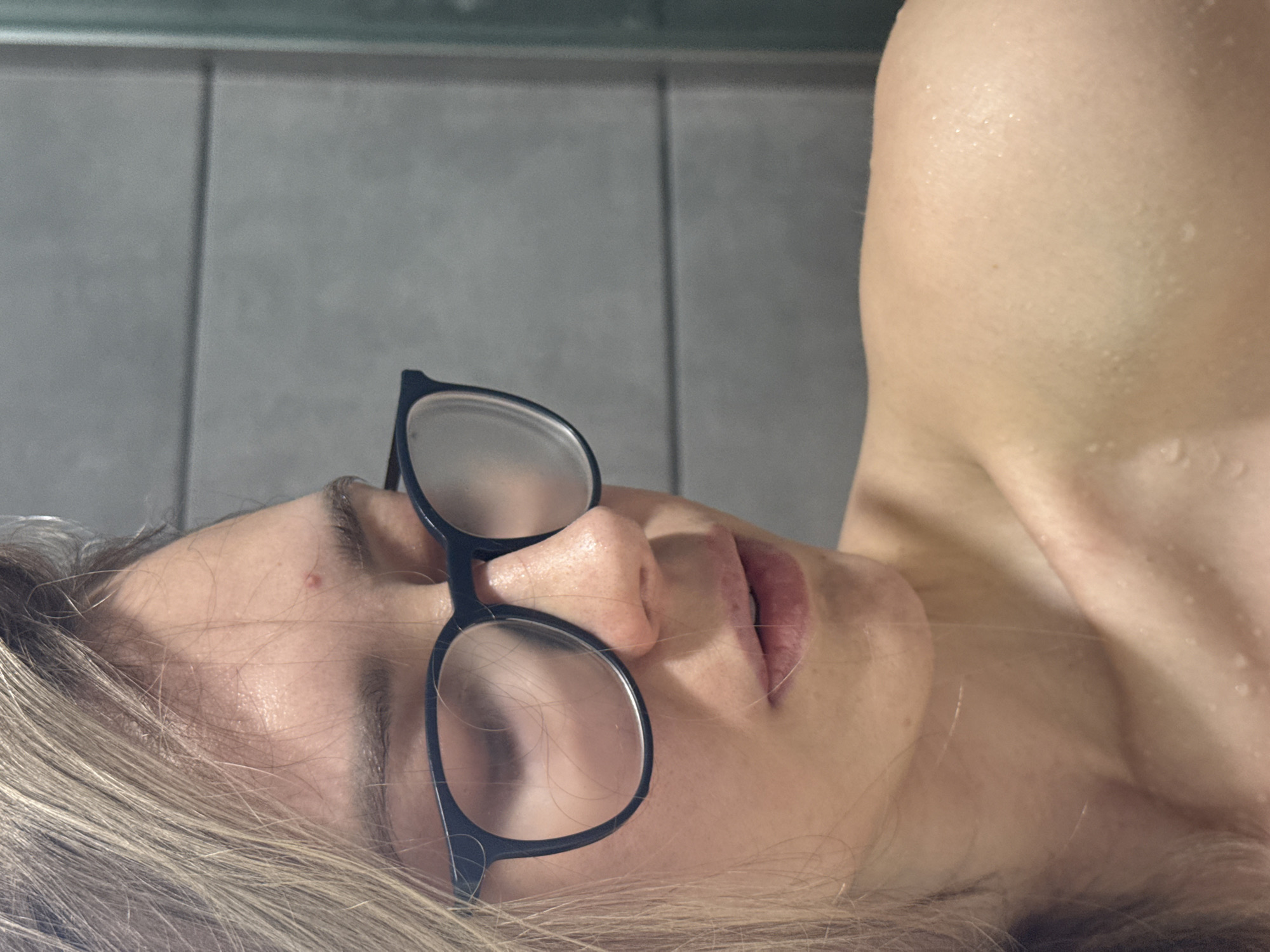 A close-up, tilted image depicts a fair-skinned person with wet skin lying down. They have light blonde hair, and are wearing black-rimmed eyeglasses. The background consists of gray tiles. No specific location in Chaikovsky, Russia, is identifiable from the image.