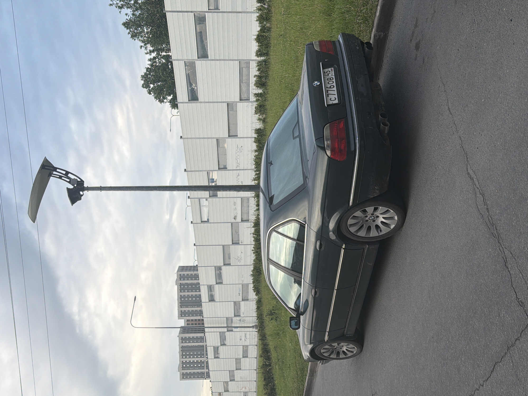 A dark-colored BMW sedan is parked on the right side of a paved road, facing left. The car is viewed from its rear quarter, showing the driver's side. The license plate reads "C776OB 45 RUS". Behind the car, a tall, modern light pole with a large, curved fixture stands near a wide, white noise barrier wall. The wall has multiple rectangular sections, some with visible graffiti. Beyond the wall, several tall, multi-story apartment buildings are visible against a cloudy sky. Lush green grass and vegetation line the right edge of the road and extend behind the noise barrier. The road surface shows some cracking.