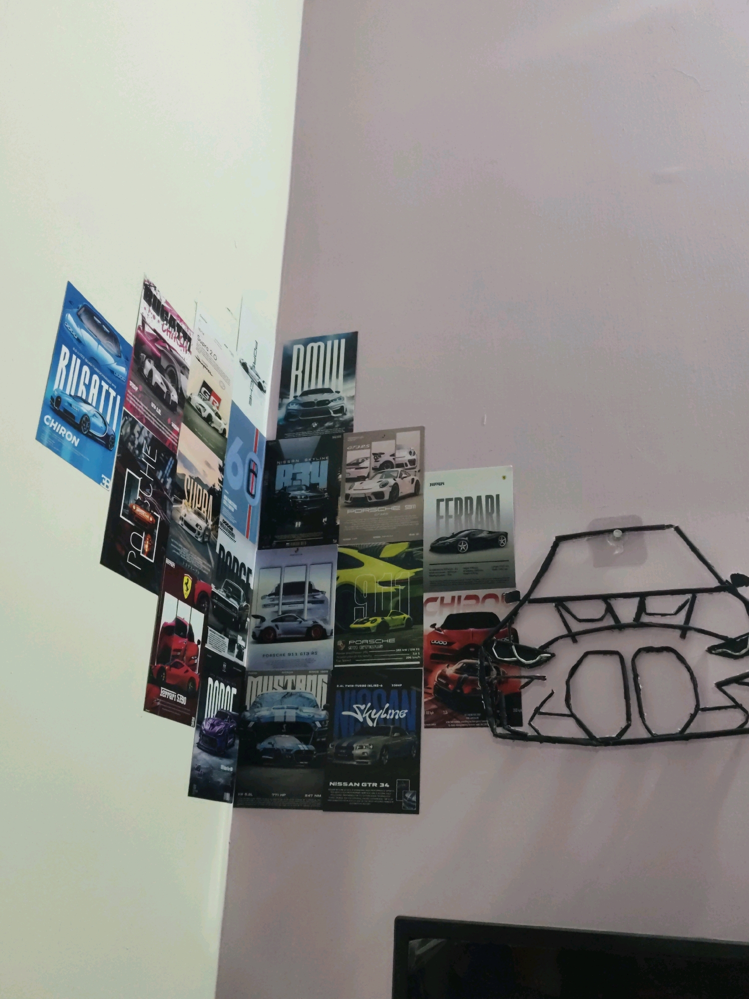 The image depicts a wall adorned with various car posters, predominantly featuring sports cars and high-performance vehicles. These posters are arranged in a somewhat scattered, overlapping fashion on a light-colored wall. Brands like Bugatti, Ferrari, Dodge, Porsche, and Nissan are visible on the posters. In the foreground, to the right, a black wireframe outline of a car's silhouette is attached to the wall, creating a decorative element. The overall scene suggests a personal collection or a themed decoration focused on automobiles. The location is identified as Sankalapura, India.
