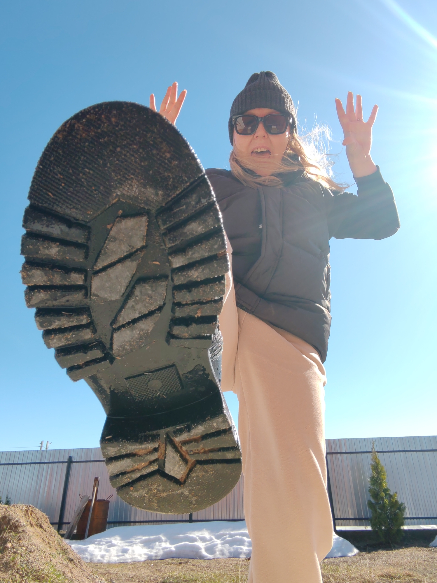 A low-angle, close-up shot depicts a woman wearing a dark beanie, sunglasses, and a black puffer jacket over beige pants, extending her leg toward the camera, revealing the sole of her dark boot. Her hands are raised with fingers spread. The sky is visible as a light blue expanse, with a bright light source causing lens flare. The background shows a fence, a mound of dirt, patches of snow, some gardening tools (a shovel and wooden planks), and a small evergreen tree. The image was taken outdoors in what appears to be a residential area in the city of Chaikovsky, Russia.