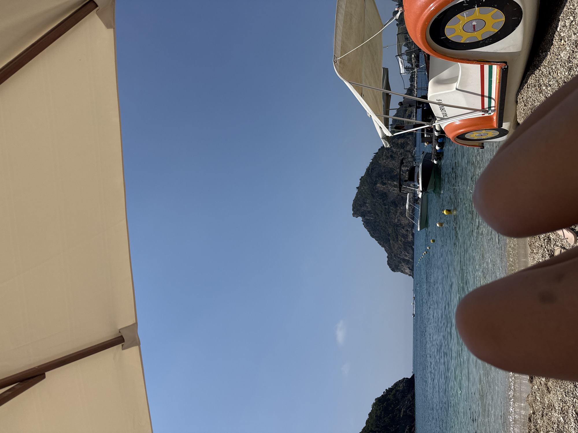 The image captures a beach scene in Palaiokastritsa, Greece, on a clear, sunny day. In the foreground, a person's legs are visible, suggesting they are lying on the pebble beach. An open beach umbrella, casting a shadow, is positioned overhead, framing the top of the image. To the right, a distinctive orange and white pedal boat, designed to resemble a car with the text "TAXI DRIVER 6", is parked on the shore. The clear turquoise water of the sea stretches out, with buoys marking a swimming area. In the background, two boats are anchored offshore, and the distinctive mountainous coastline of Palaiokastritsa, with lush green vegetation, rises from the water. The sky is a vivid blue, with a single small cloud visible. The overall impression is one of a tranquil summer day at a picturesque coastal location.