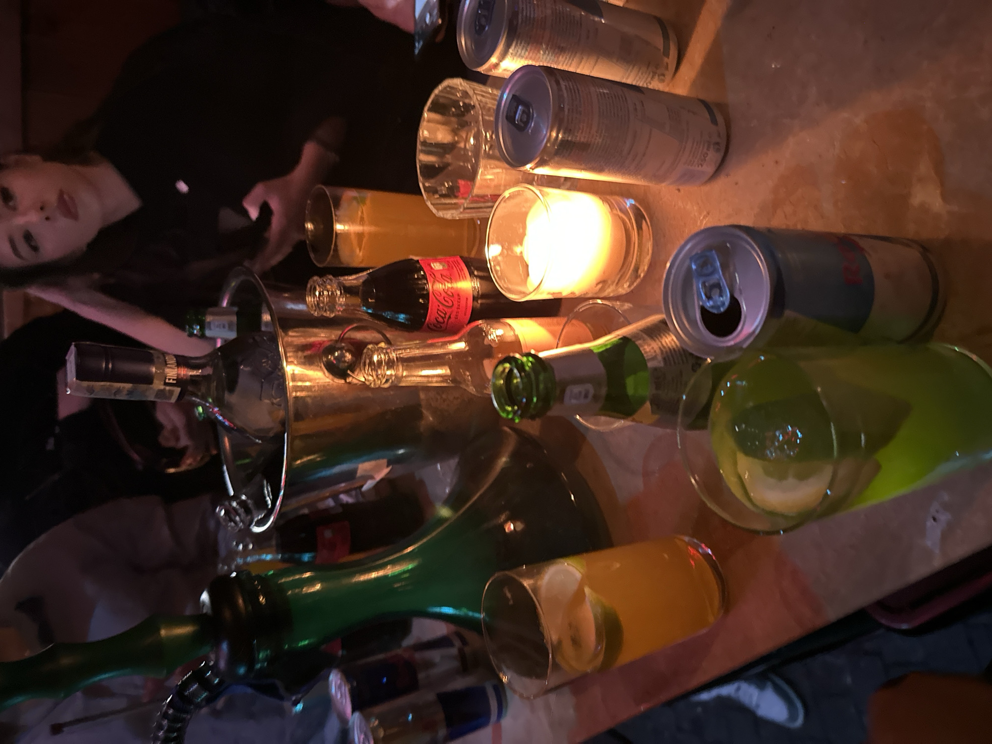 Scene: An indoor bar or club setting, Plovdiv, Bulgaria.
People: A woman with long dark hair and fair skin is visible, partially obscured by objects on the table. Another person is partially visible, obscured by the hookah.
Subjects: A hookah, likely in use.
Objects: An ice bucket with liquor bottles, including what appears to be Finlandia Vodka. A small Coca-Cola bottle is also visible. There are several opened cans of Red Bull energy drink scattered around. Various glassware including stemmed glasses and short cylindrical glasses are present, some containing liquid. A lit candle is also present on the table.
Actions: The environment suggests a social gathering, possibly involving consumption of alcohol and hookah smoking.
Interactions: The objects are arranged to facilitate drinking and smoking hookah.
Environment: The location appears to be dimly lit and crowded, suggestive of a bar or club atmosphere. The table surface is light-colored and reflective.