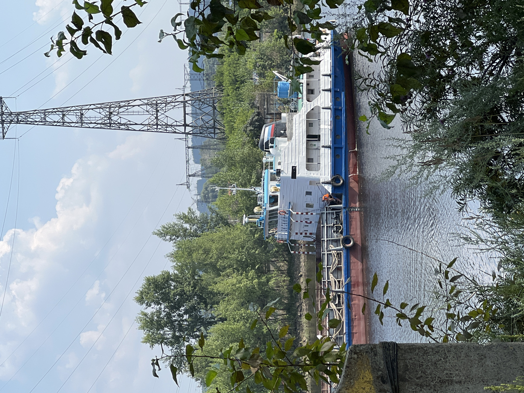The image captures a view of the Kama River in Chaikovsky, Russia, on a sunny day with scattered clouds. A white and blue river vessel, possibly a tugboat or a small cargo ship, is docked or moving slowly on the water. The boat has multiple windows and railings, with red and blue accents along its hull. In the background, a tall, industrial power transmission tower stands prominently against the sky, surrounded by a dense forest of green trees. The foreground is partially obscured by foliage and a weathered concrete pillar. The water appears calm with ripples.