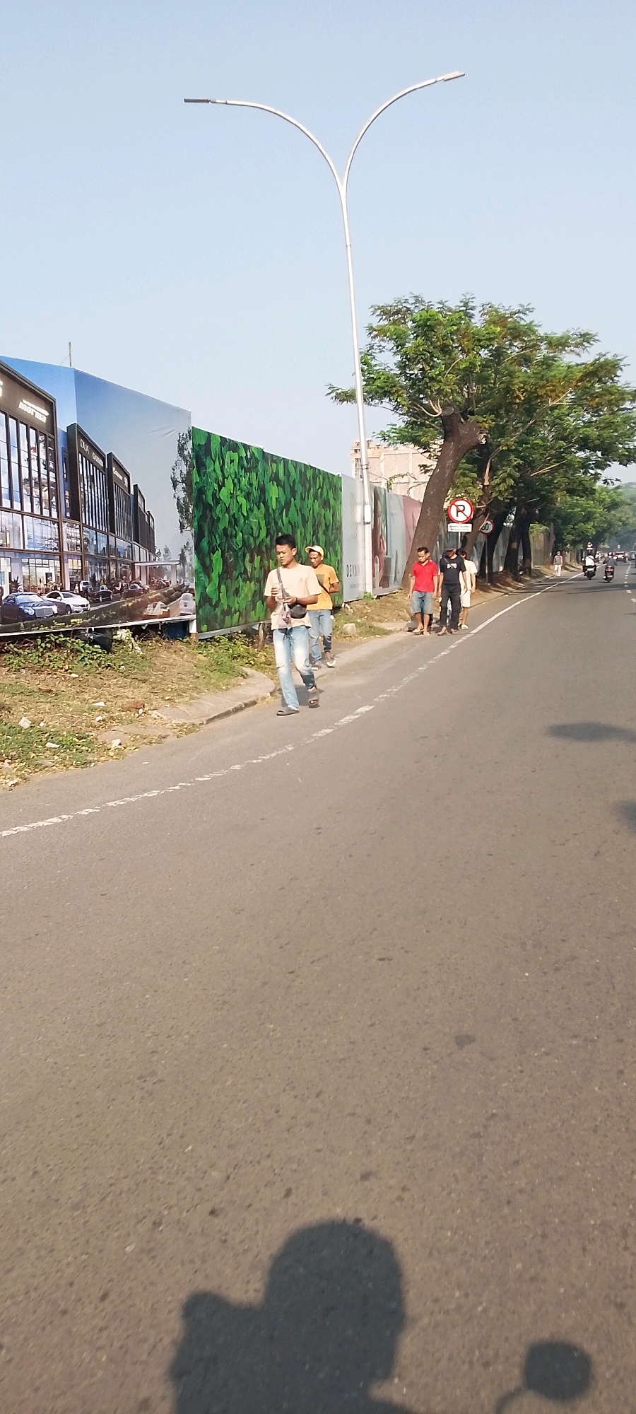 The image depicts a street scene in Gunung Putri, Indonesia, on a clear day with bright sunlight casting shadows. In the foreground, the asphalt road stretches into the distance, with a white dashed line marking the lane. On the left side of the road, a large, colorful billboard displays an architectural rendering of a modern building complex and lush green foliage. Adjacent to the billboard is a patch of dry grass and vegetation.
Several individuals are walking along the right side of the road. The most prominent is a young man in a light-colored t-shirt and blue jeans, carrying a bag across his chest. Behind him, another person in a yellow shirt is walking, followed by two more individuals dressed in a red shirt and black shirt, respectively. Further down the road, more people are visible, and a motorcycle is seen in the distance.
To the right of the road, a line of trees with dense green canopies provides shade. A street lamp with a modern, curved design stands tall against the sky. A red circular traffic sign with a white horizontal bar and a "P" symbol is mounted on a pole, indicating a parking restriction. The environment appears to be an urban or semi-urban area undergoing construction or development, suggested by the large billboard and the general road conditions. The overall atmosphere is one of everyday life in a developing Indonesian city.