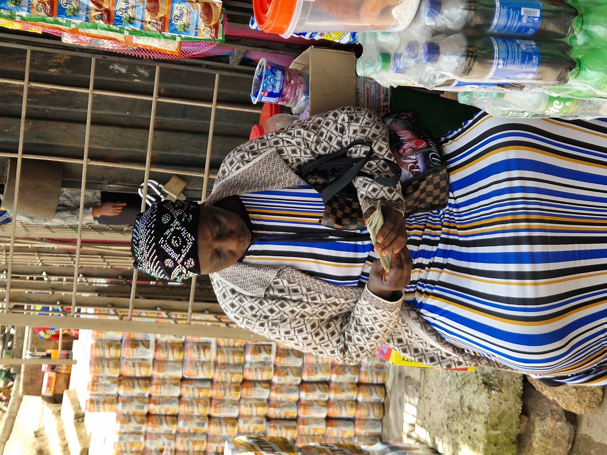 In Kabong, Nigeria, a woman is pictured at an outdoor market stall. She wears a black and white headscarf and a patterned jacket over a boldly striped dress, the colors of which include blue, white, yellow, and black. Her attention is focused downward as she counts a small wad of cash in her hands, suggesting a transaction. Behind her, a metal grate forms the backdrop of the stall, behind which are stacked cans and other goods. To her right, a variety of items are displayed for sale, including small packets of food products and several plastic bottles of beverages. The scene is illuminated by natural daylight, casting shadows and highlighting the textures of the various items and the woman's attire.