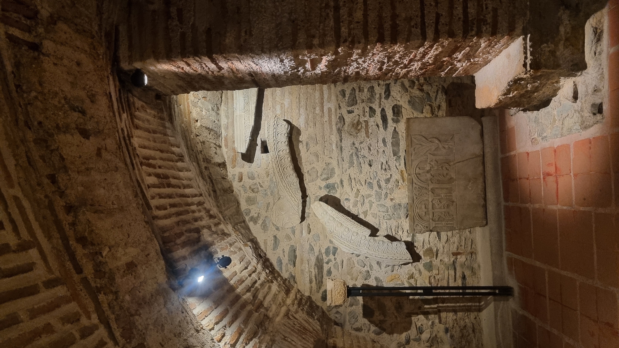 The image shows an interior view of a dimly lit, ancient stone room, likely in Thessaloniki, Greece, given the context of a historical excavation or museum display. The room features a vaulted ceiling constructed of bricks and stone, with exposed rough stone walls. Several ancient stone artifacts are displayed: a large, rectangular stone slab with intricate carvings of religious iconography, likely a sarcophagus lid or altar piece, is positioned prominently in the foreground. Above and to the left of this slab, two large, curved stone fragments with decorative carvings are mounted on the wall. To the left of the wall carvings, a stone pedestal with a small, bowl-like top stands on a dark metal pole. A rough-hewn stone bench is visible to the right, against a brick pillar. The floor is paved with reddish-brown square tiles. Artificial lighting from spotlights illuminates the artifacts and the textured surfaces of the room. There are no people visible in the scene.