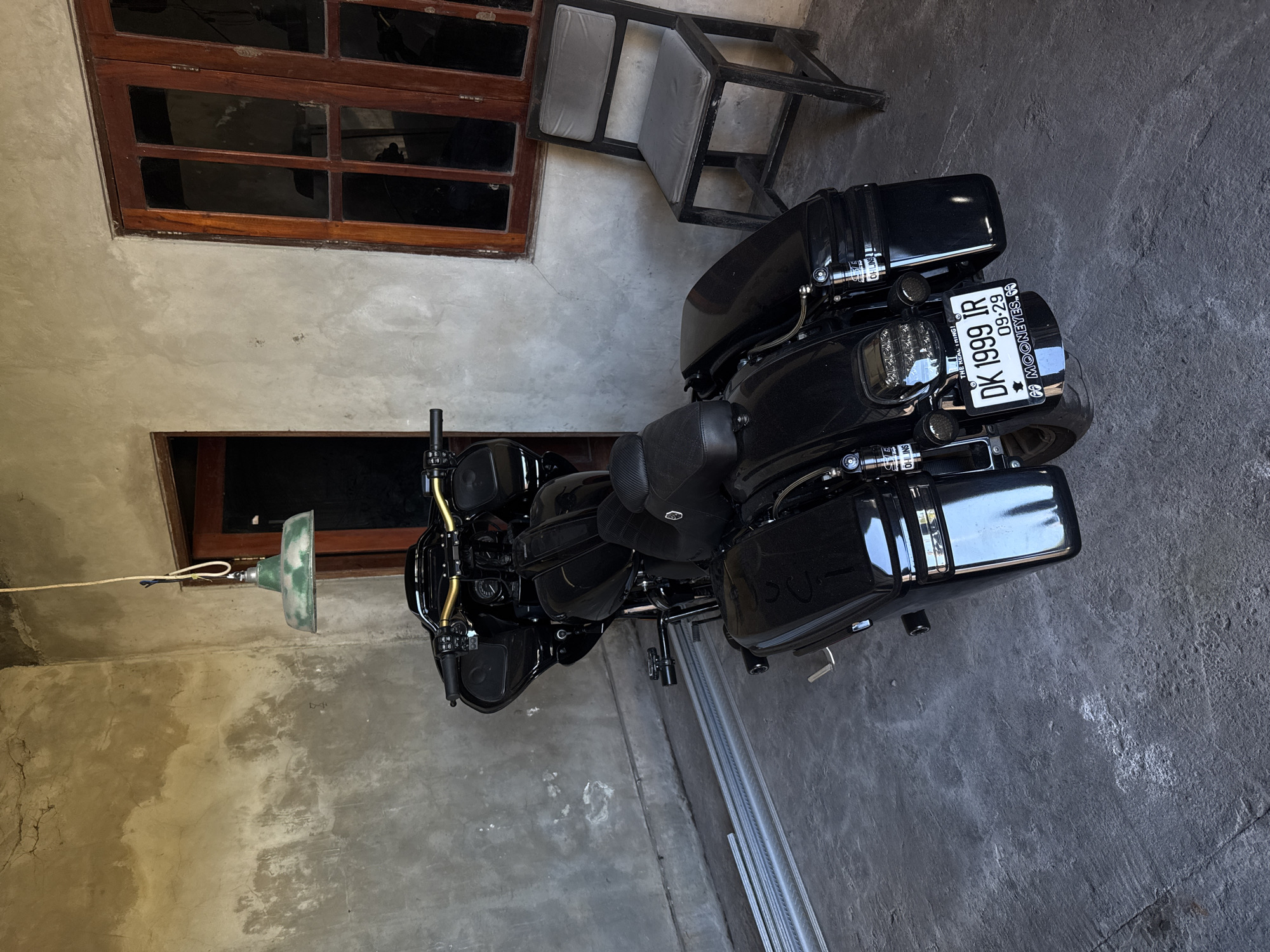 The image depicts a black motorcycle parked indoors. The license plate reads "DK 1999 IR 09-29", indicating it's registered in Denpasar, Bali, Indonesia. The interior setting appears to be a garage or workshop, with exposed concrete walls. To the left, there is a ceiling lamp, hanging by a cord. There are window frames with partially obscured glass in the background. A grey metal step stool stands near the motorcycle. Based on the license plate, the motorcycle is likely in Denpasar, Indonesia; however, the specific location of the North Kuta is not discernible from the image itself.