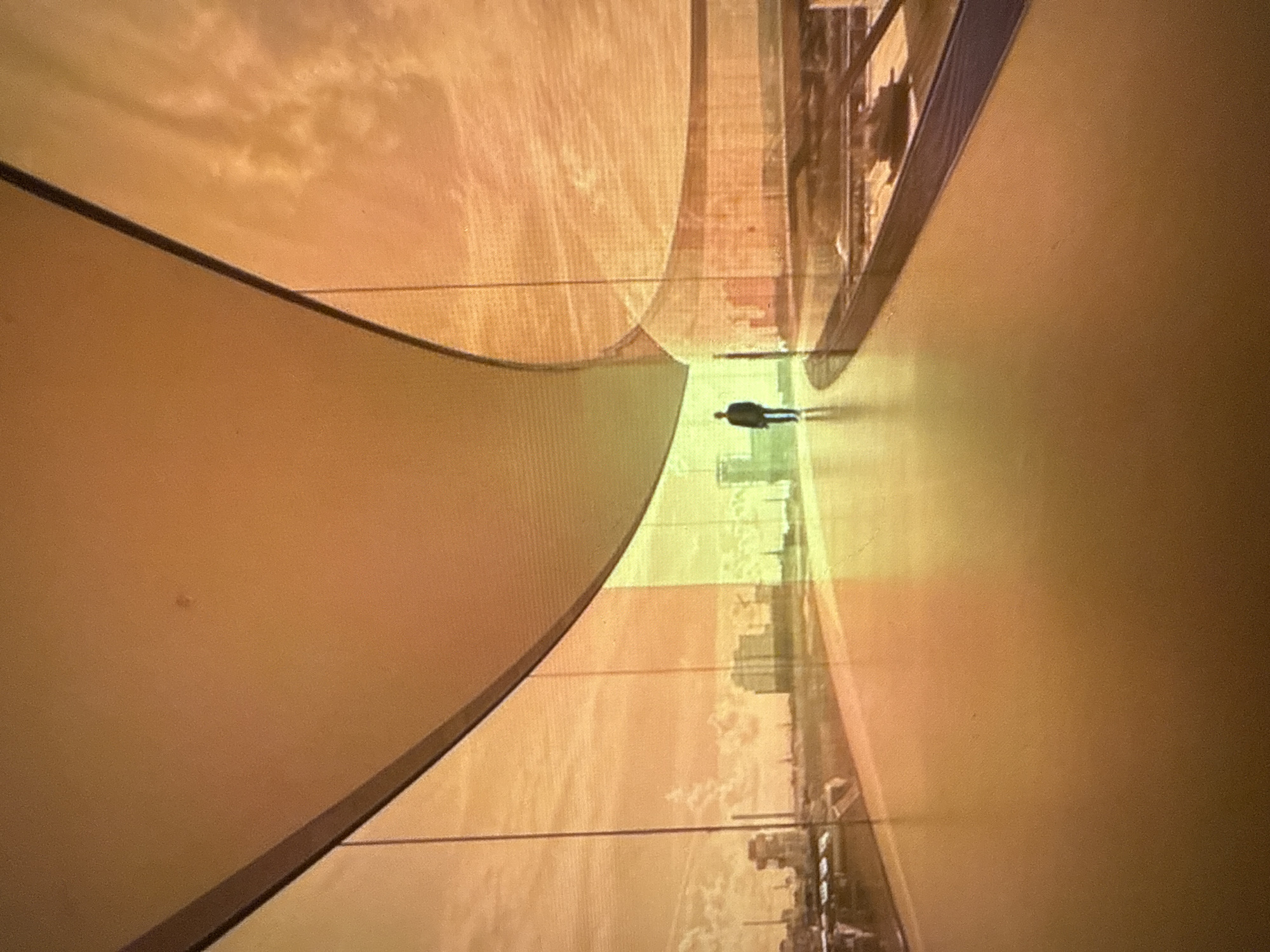 The image shows a person walking along a curved walkway with large glass windows. The windows offer a view of a city skyline with buildings and industrial structures. The sky appears to be a warm, orange hue, suggesting either a sunset or sunrise. The walkway is made of a polished, light-colored material that reflects the surrounding environment. The overall impression is one of modern architecture and urban landscape. The image does not contain any information that would allow for the identification of the city of Boreti, Montenegro.