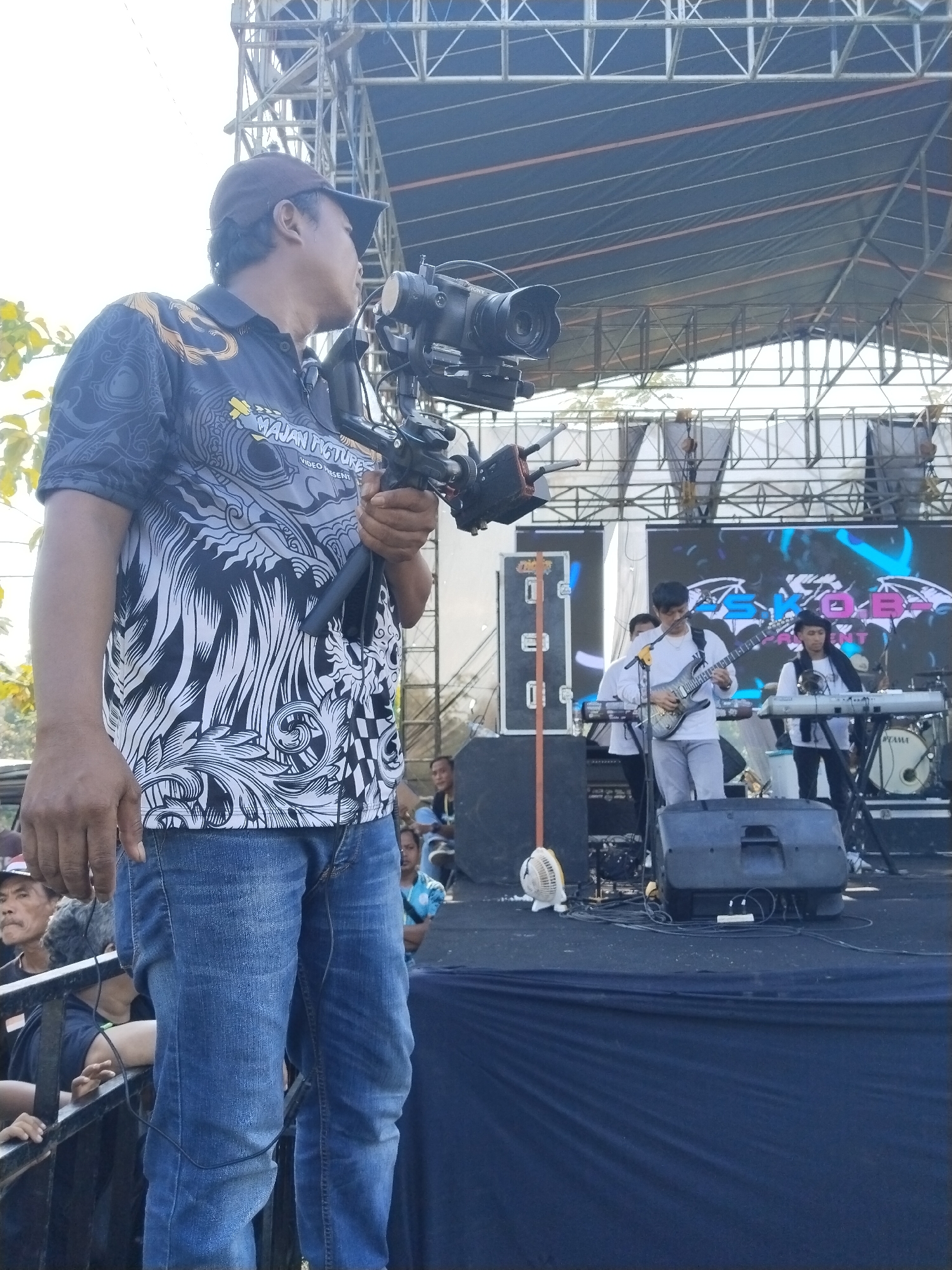 A cameraman stands in front of a stage in Karanganom, Indonesia. He is wearing a brown cap, a patterned black and white shirt, and blue jeans. He is holding a professional video camera mounted on a stabilizer rig. Behind him, on the stage, a band is performing with a guitarist, a keyboardist, and other members partially visible. There are speakers, monitors, and a drum set on the stage. A crowd is visible in front of the stage behind a metal barrier. The stage is under a metal frame structure.