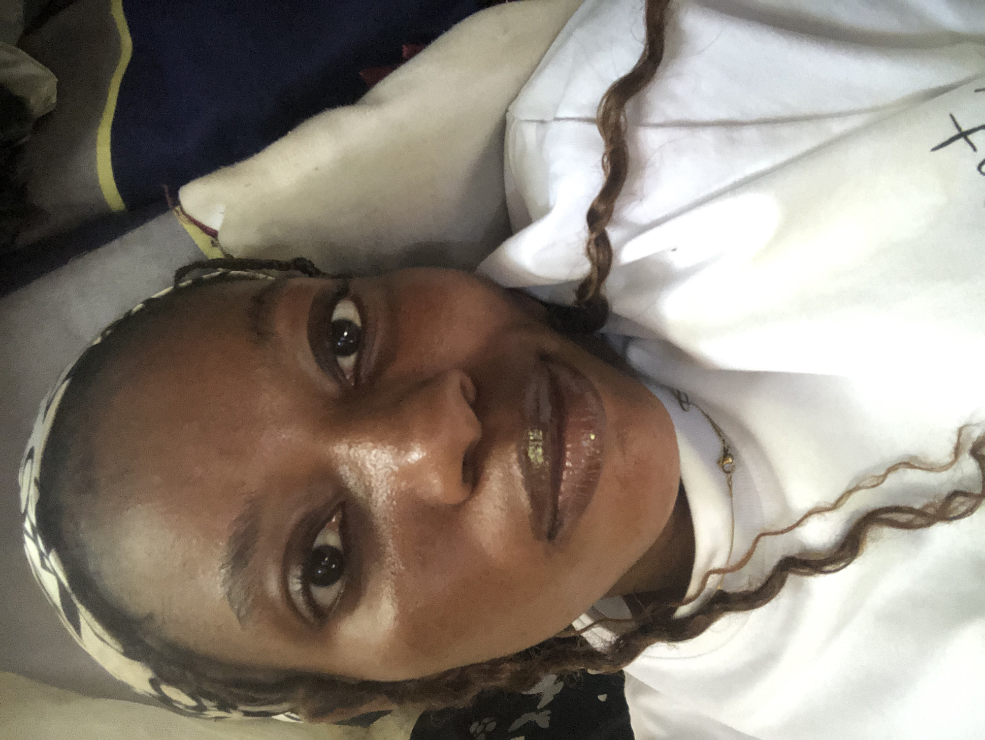 A close-up, overhead shot of a young Black woman lying down, with her head tilted and facing the camera. She has long, brown, curly hair, some of which falls across her face and chest. She is wearing a white t-shirt and a patterned headwrap. The background consists of bedding and a pillow. The image appears to be taken indoors, likely in a bedroom, in Pindiga, Nigeria.
