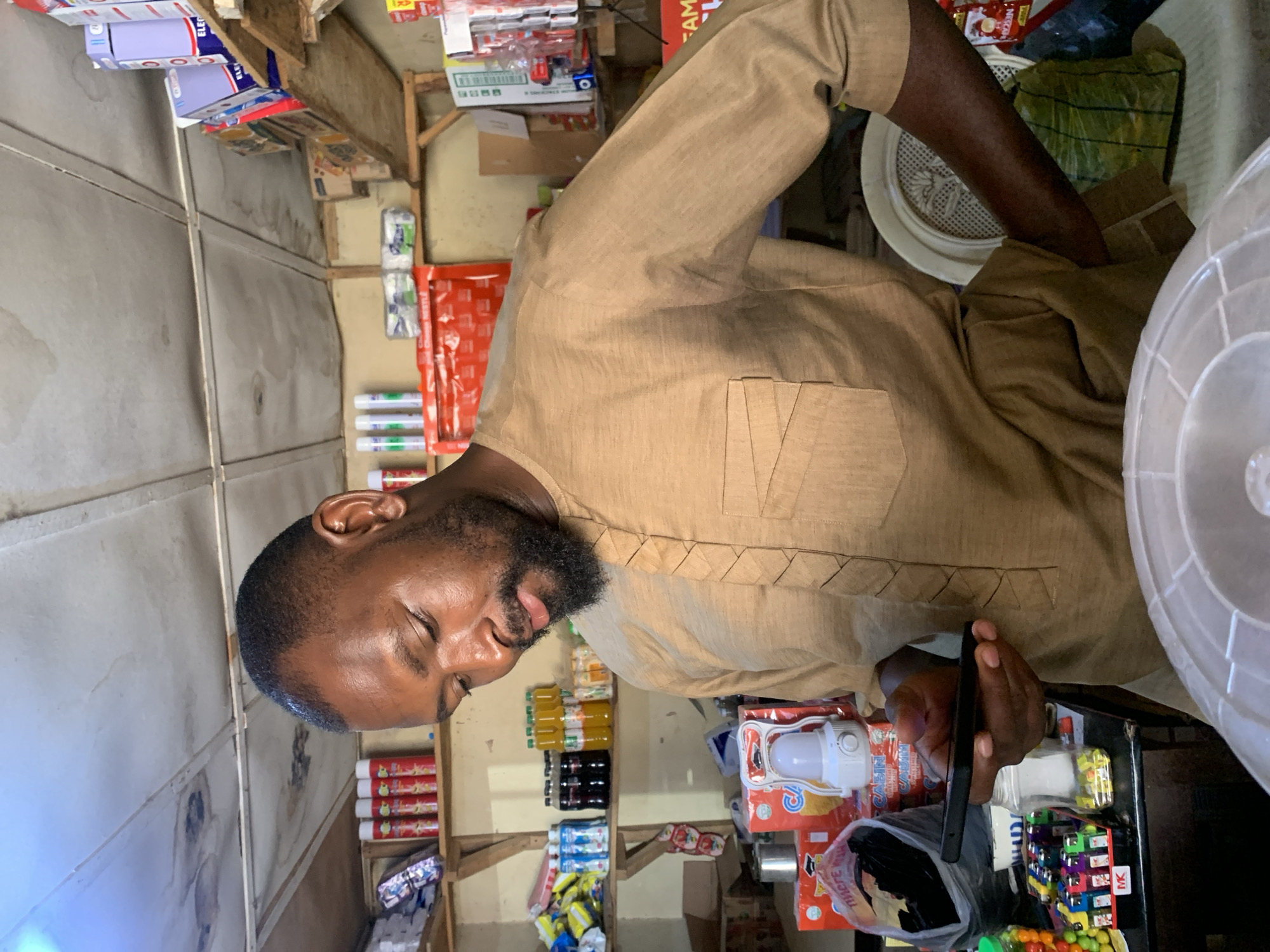 An adult man with short, dark hair and a beard is standing inside a small shop. He is wearing a light brown shirt and holding a black smartphone in his left hand. The shop is stocked with various goods, including food items, beverages, and household products, arranged on shelves. The shop appears to be in Jos, Nigeria.
