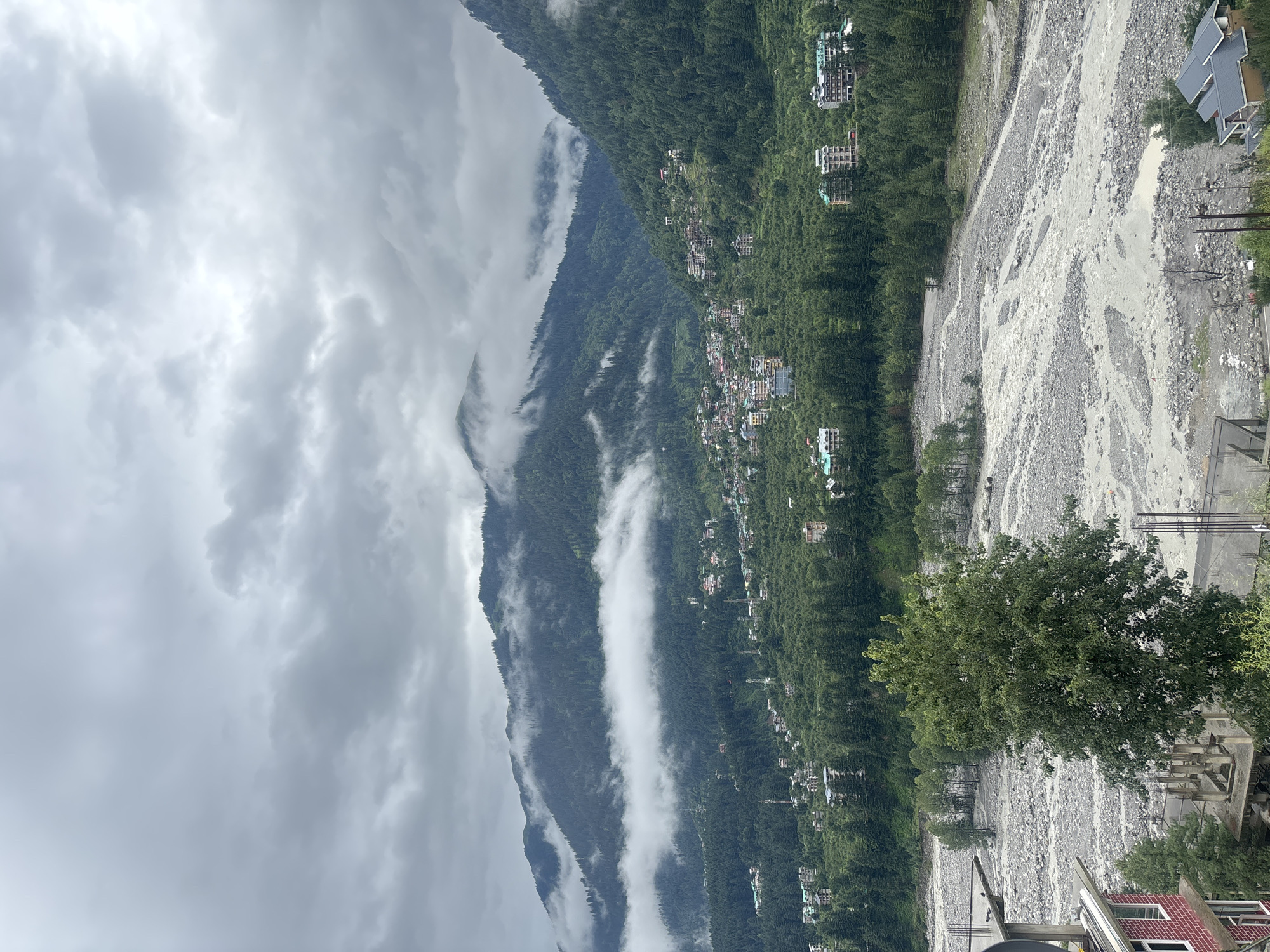 This is a high-angle, wide shot of a mountainous region near Vashisht, India. The sky is overcast with heavy gray clouds, casting a muted light on the landscape. In the foreground, a wide, gravelly riverbed dominates the lower portion of the image, with a narrow, murky stream flowing through it. A large, leafy green tree stands in the middle ground, to the left of the riverbed, obscuring some of the view.
Behind the riverbed, a town is nestled on the slopes of the surrounding mountains. Buildings of varying sizes and architectural styles, some appearing to be hotels or residences, are scattered amongst dense green pine forests. The mountains themselves are covered in lush vegetation, with patches of white clouds clinging to their peaks and slopes, creating a dramatic, misty effect. Several cell towers are visible throughout the town and on the mountainside. In the far left foreground, the corner of a red brick building is visible, suggesting the presence of more development. The overall scene depicts a rural, mountainous area with a settled community integrated into the natural environment.