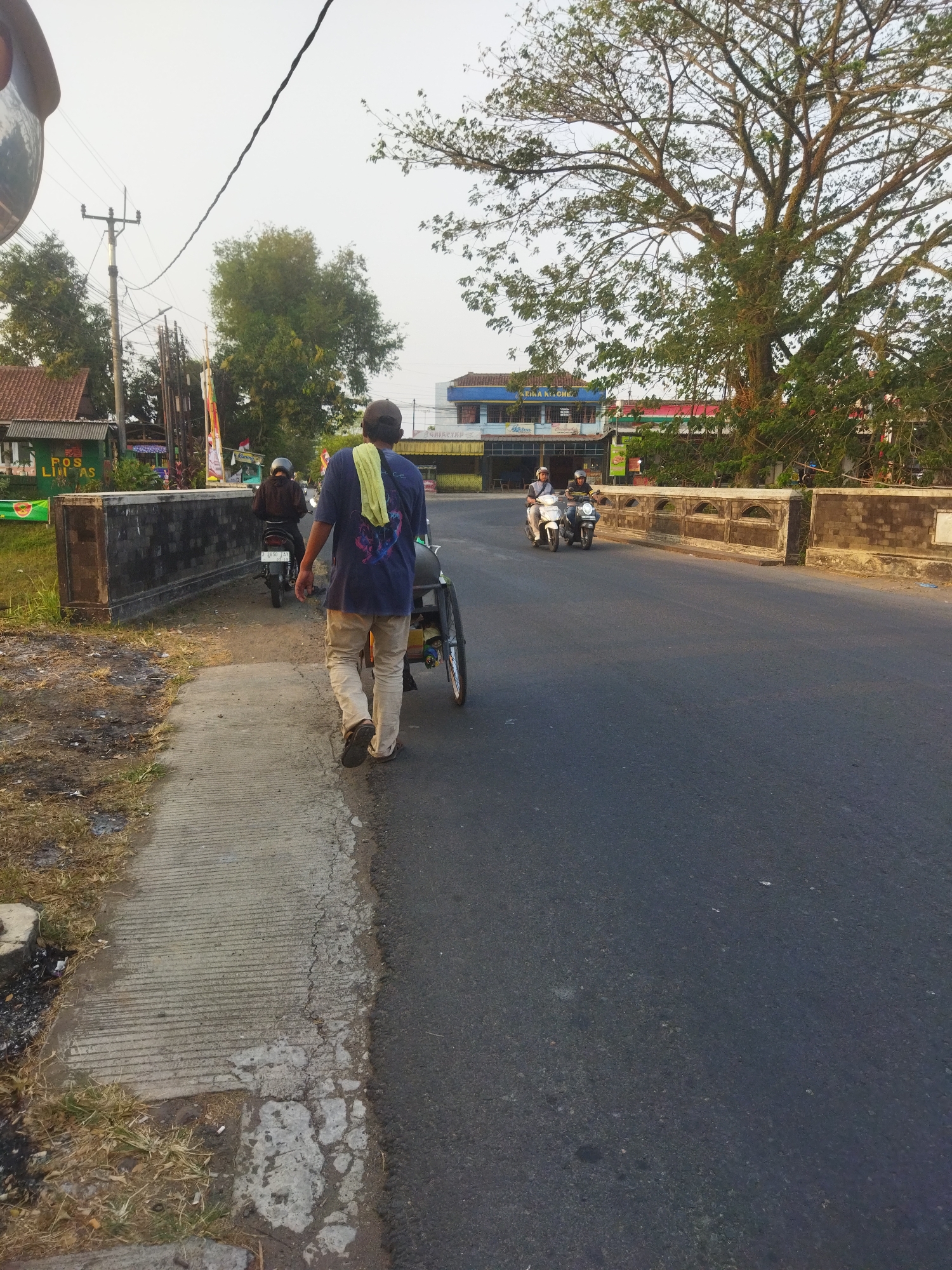 In Banjar, Indonesia, a man walks along a concrete sidewalk next to a paved road. He is wearing a blue t-shirt and tan pants, with a yellow cloth draped over his shoulder. He is pulling a three-wheeled cart behind him. Two motorcycles are visible in the distance on the road, with riders wearing helmets. A bridge with a stone railing stretches across the road in the background. Buildings and trees line the sides of the road, under a hazy sky. The scene appears to be a street or intersection in a semi-urban area.