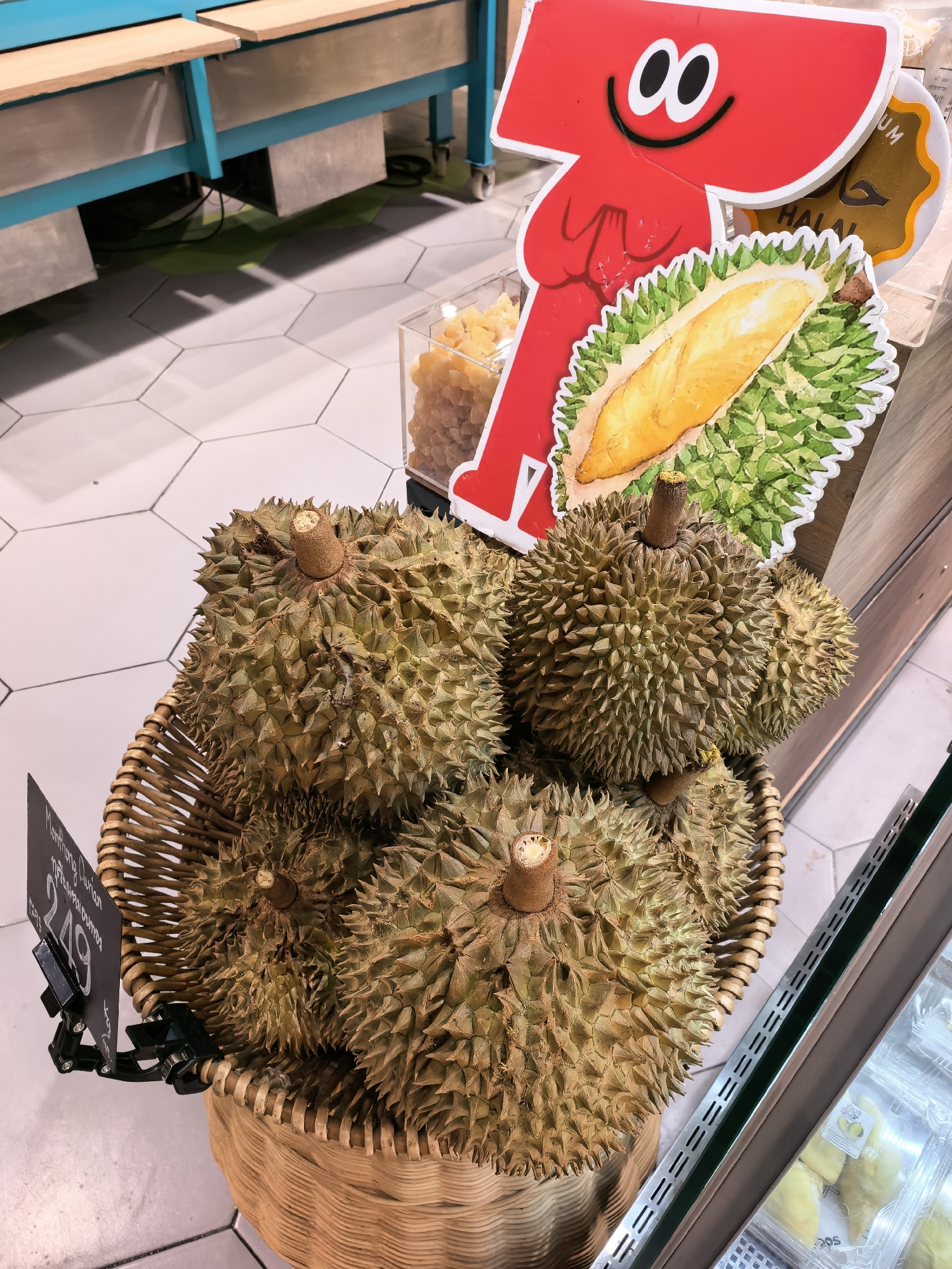 A close-up, high-angle shot shows a woven basket overflowing with durians, a tropical fruit known for its distinctive thorny rind and strong odor. The durians are a light brown and green color, with sharp spikes covering their surfaces. Some of the durians have their stems visible. A price tag, black with white text and a black clip, is attached to the front of the basket, partially obscuring the durians. The text on the tag appears to be in English and possibly another language, with the price "249" prominently displayed. Behind the basket, a colorful red cartoon cutout of a durian segment with a smiling face is visible, alongside other signage. The floor is tiled in a hexagonal pattern, with some light and dark gray tiles. In the background, a blue metal structure with wooden shelves suggests a market or retail setting. Further to the right, a refrigerated display case with clear plastic containers holding yellow fruit segments can be seen. The overall scene suggests a fruit stand or market in a tropical location, potentially Ko Samui District, Thailand, given the presence of durian and the implied climate.
