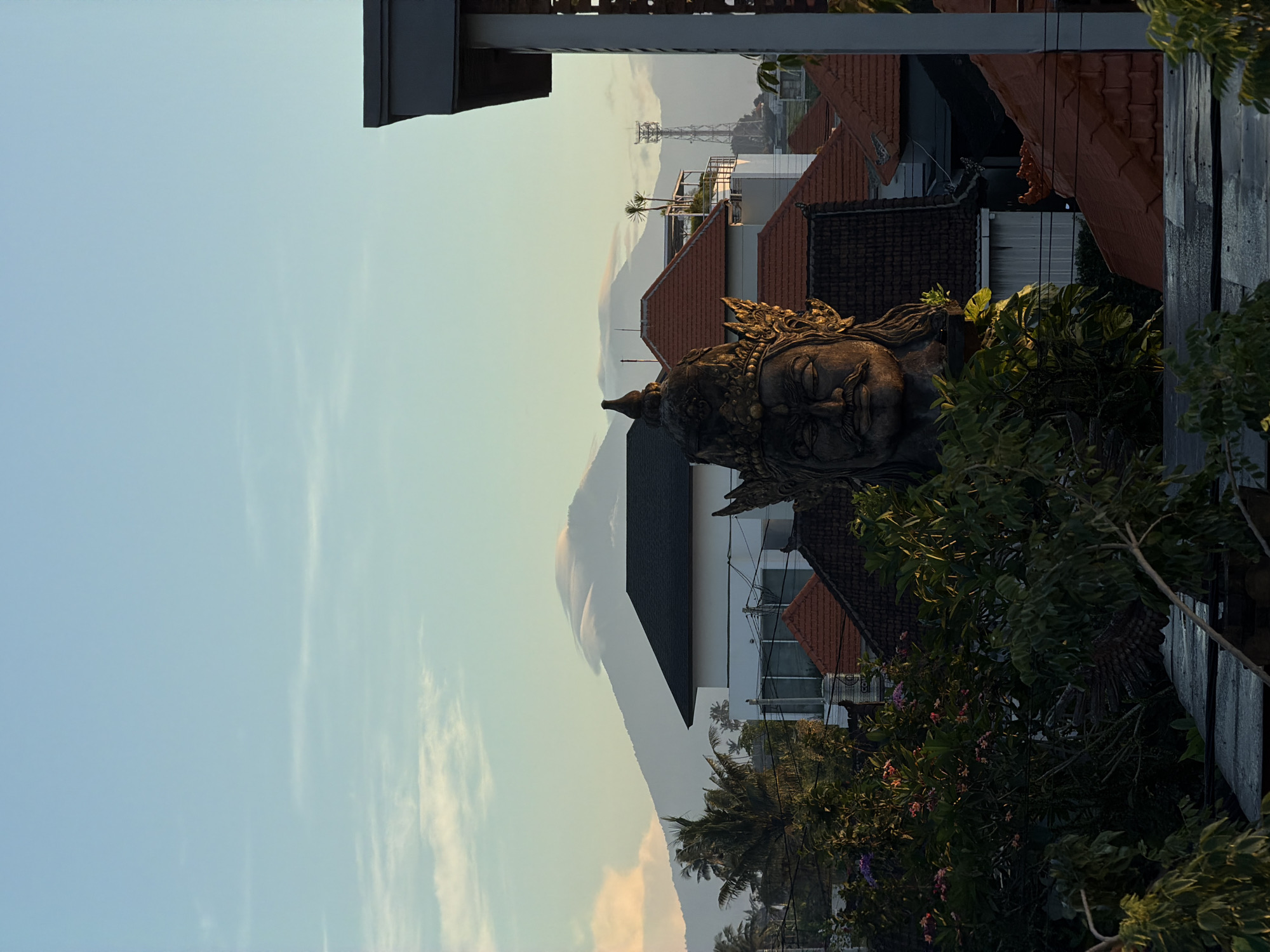 The image shows a serene view in Mengwi, Indonesia. A stone statue of a deity, possibly Buddha, with intricate headwear, is nestled amongst green foliage, including trees with flowering purple blossoms. Behind the statue, a landscape unfolds with buildings, including those with red-tiled roofs and white walls, and mountains fading into the horizon. A communication tower is faintly visible. The sky transitions from light blue to a warm yellow-orange hue, suggesting either sunrise or sunset. The image is oriented vertically.
