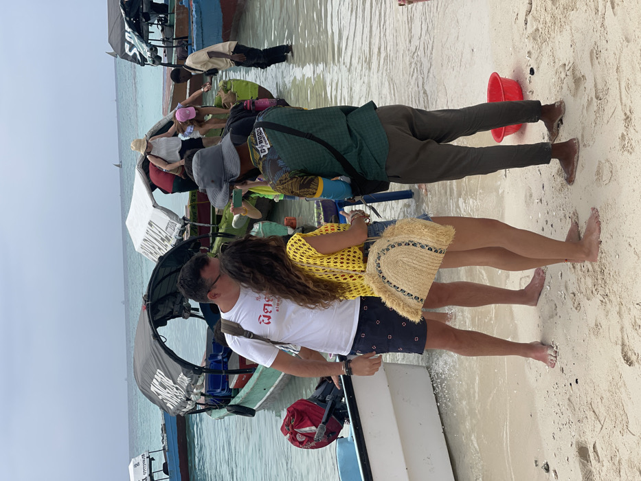 The image depicts a busy beach scene in the city of Zanzibar, Tanzania, under an overcast sky.
In the immediate foreground, three individuals are standing barefoot. From left to right: a Caucasian male, approximately 30-40 years old, wearing dark sunglasses, a white t-shirt with red script, dark patterned swim shorts, and a dark backpack, stands in shallow water. Next to him is a Caucasian female, 20-30 years old, with long brown hair, wearing a yellow open-knit sleeveless top, light-colored shorts, and carrying a fringed, semi-circular straw bag. To their right, an African male, 20-40 years old, wears a grey bucket hat, a patterned green t-shirt with "G+BOOST" text, dark trousers, and a black shoulder bag, positioned on drier sand. A red plastic bucket rests on the sand near his right foot.
The midground is occupied by multiple small, motorized wooden boats partially beached in the clear, turquoise water. A green boat with a black canopy displaying text (partially legible as "SAKURA") is central, with a blue/red boat featuring a white canopy to its right. Several individuals are engaged in boarding or disembarking these vessels; a Caucasian female in a straw hat and striped top is visible stepping from the green boat, while an African male stands in the water, appearing to assist. A smaller, light blue boat with an outboard motor is partially visible on the far left. The background shows a calm sea with several distant sailboats on the horizon.