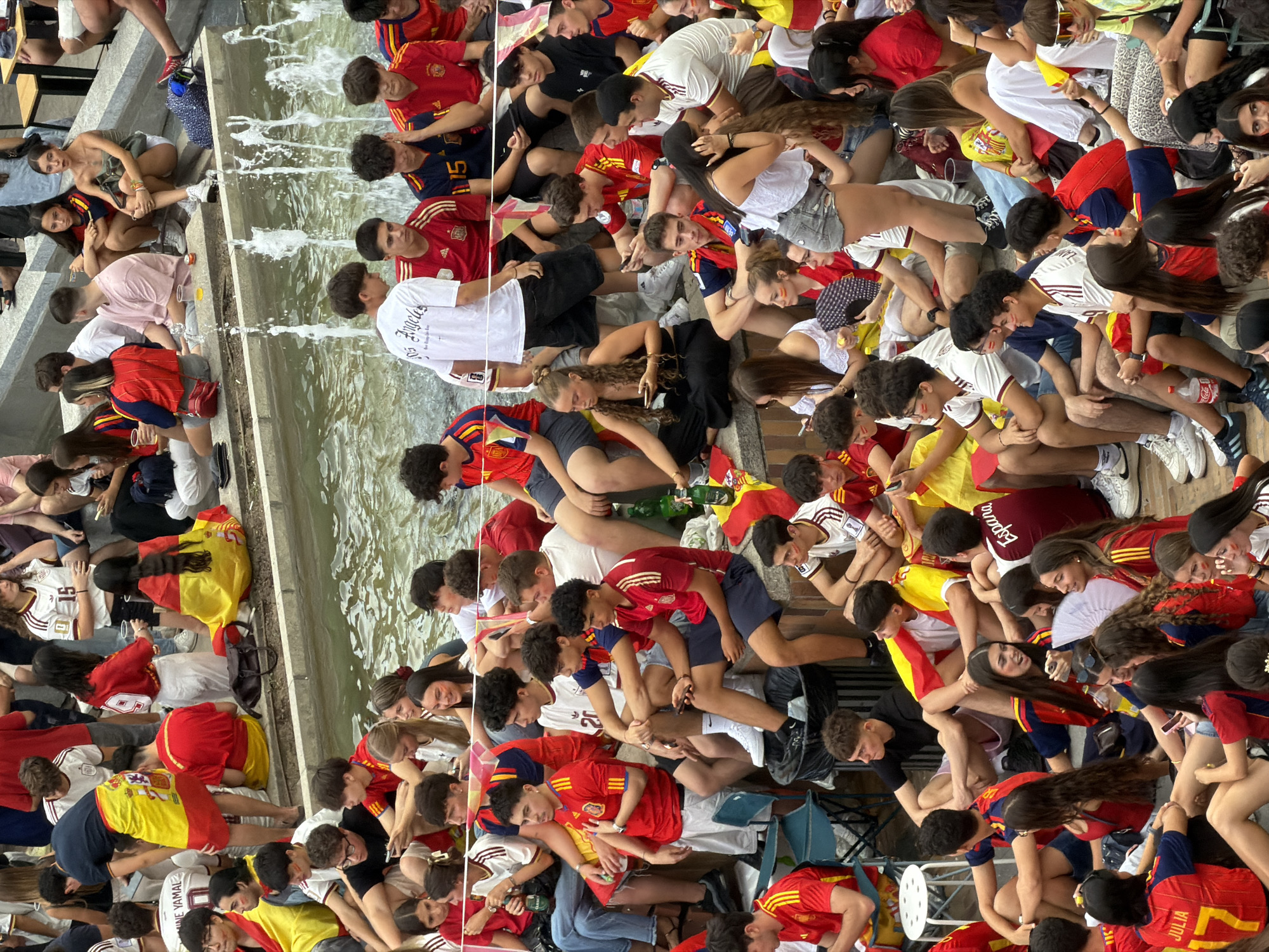 The image depicts a crowd of people gathered near a fountain, likely watching a sporting event or celebration. Many individuals are wearing red and yellow attire, suggesting a Spanish national team or event. Some are holding Spanish flags and scarves. The setting appears to be an outdoor urban space, possibly a public plaza or park. The fountain is spraying water, indicating it's active. People are sitting, standing, and interacting with each other, with some using mobile phones. The overall atmosphere seems lively and engaged.
Given the provided information, it is not possible to ascertain the specific location within the city of Las Rozas de Madrid. The image itself does not contain any recognizable landmarks or signage that would pinpoint a particular spot.