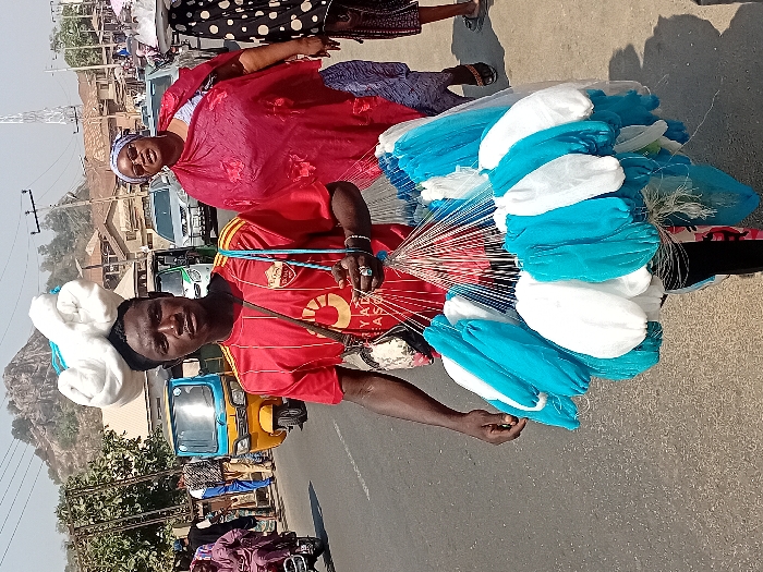 A man in a red soccer jersey stands on a street carrying a large bundle of blue and white netting. A red-dressed woman stands nearby. He has some netting piled on his head. In the background, there is a yellow auto-rickshaw and various buildings and a hill. The netting is mostly white and blue, but some of the netting has silver.
