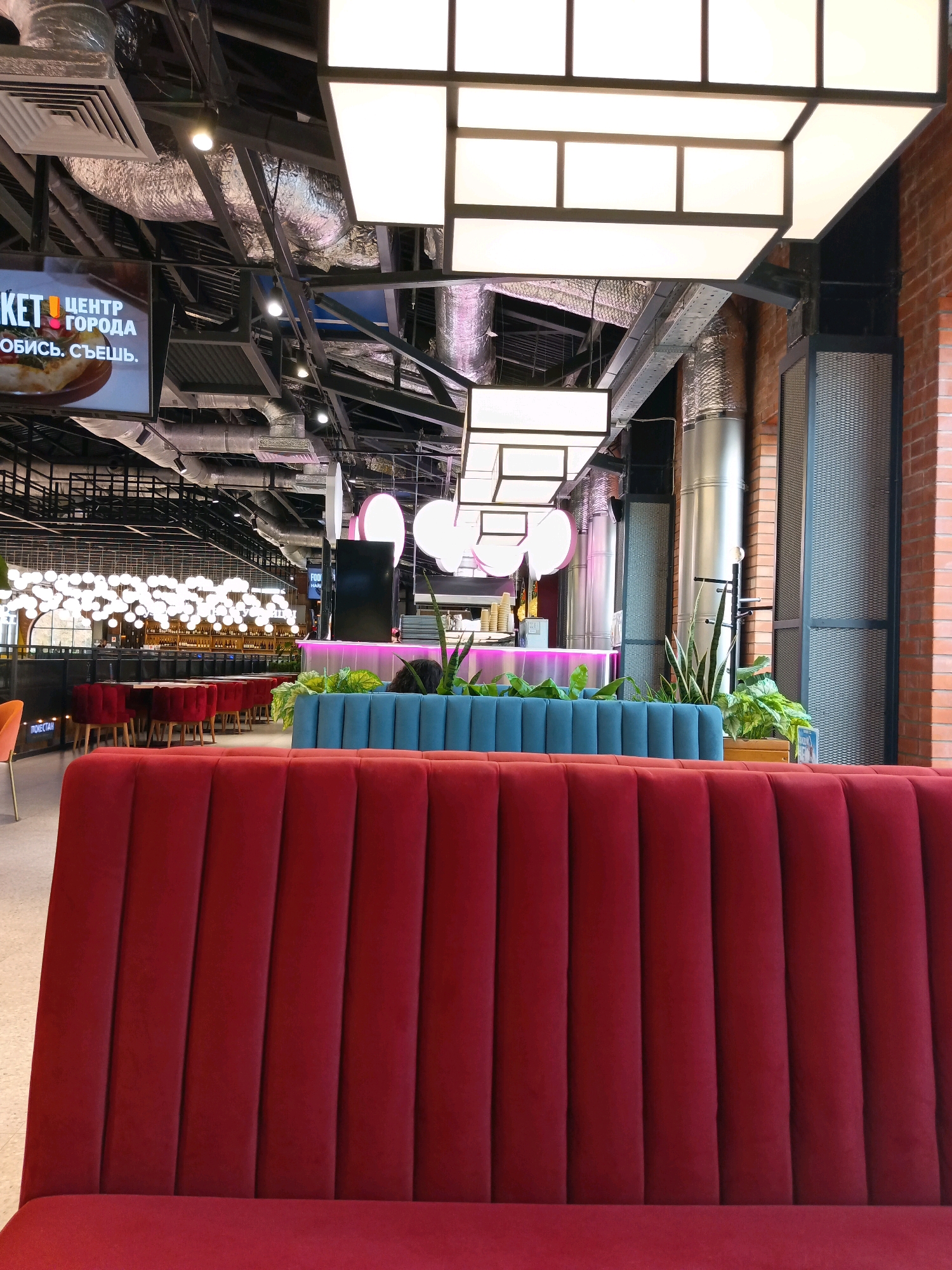 The image depicts the interior of a modern food court or restaurant, likely located in Krasnodar, Russia, given the Cyrillic text visible on a screen. The scene is characterized by a multi-level ceiling with exposed industrial elements like ductwork and metal beams, punctuated by large, white, square lighting fixtures. A prominent red upholstered bench seat dominates the foreground, suggesting a seating area. Behind this, a teal upholstered bench and a low planter with lush green plants create a separation. In the background, rows of red chairs surround tables, leading towards a bar or counter area illuminated with pink neon lighting. A large digital screen displaying text and an image of pizza is mounted on the ceiling on the left. Various industrial-style lights, some spherical, are suspended throughout the space, contributing to the contemporary and slightly industrial aesthetic. The overall environment appears clean and well-lit, suggesting a commercial establishment. There are no visible people in the immediate foreground, but hints of activity might be present in the distance.