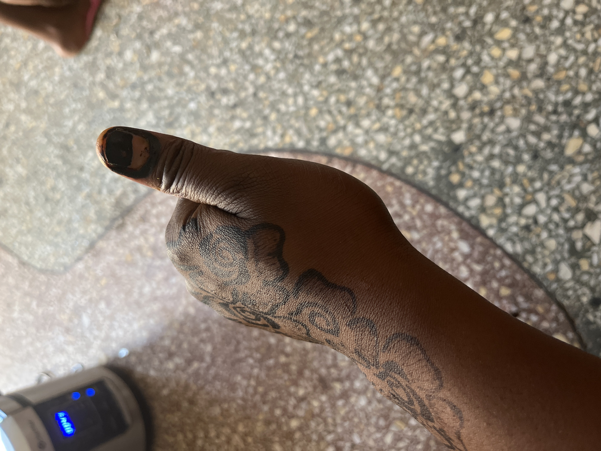 A close-up overhead shot depicts a dark-skinned human arm and hand with dark ink designs on the skin. The thumbnail is covered in dark, opaque ink. The arm is resting on a speckled surface resembling a terrazzo floor. A portion of a circular object with a reddish-brown edge is visible under the arm. In the lower left corner, a small white electronic device with blue indicator lights is partially visible. The location of the image in the city of Jos, Nigeria is not discernible from the visual information provided.