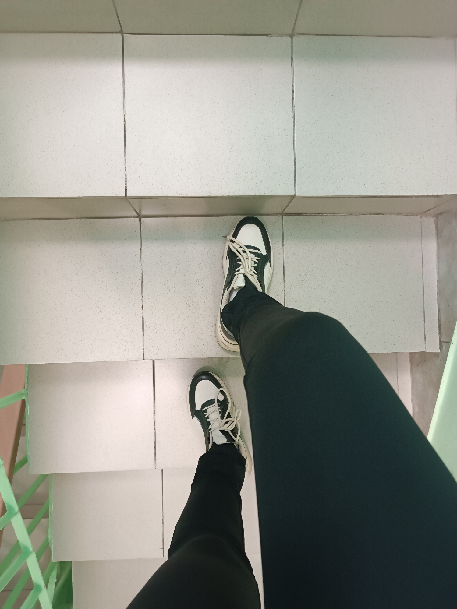 A first-person perspective photograph shows a person wearing black pants and black and white sneakers standing on white tiled stairs. A green metal railing is partially visible on the left side. The location is not discernible from the image. There is no other information present.