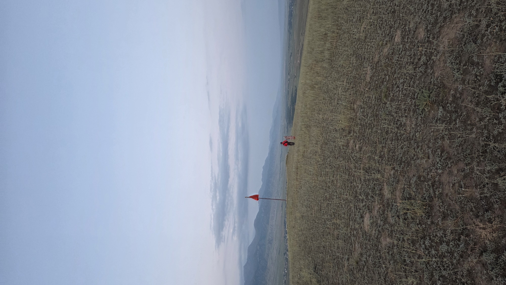 This image depicts a panoramic view of a barren, grassy hilltop overlooking a valley with distant mountains. A solitary red flag on a pole stands prominently on the hilltop. Two figures, one in a red jacket and another in a patterned outfit, are positioned near the flag, facing away from the camera and towards the valley. The sky is overcast with diffused light, suggesting either early morning or late evening. The landscape is predominantly dry and sparsely vegetated, with low-lying bushes and grasses. The valley below appears to be inhabited, with visible structures and fields stretching towards the foothills of the mountain range. The overall scene conveys a sense of isolation and natural beauty, with a hint of human presence amidst a vast, undeveloped landscape. The specific location is identified as Ак Жар, Kyrgyzstan.