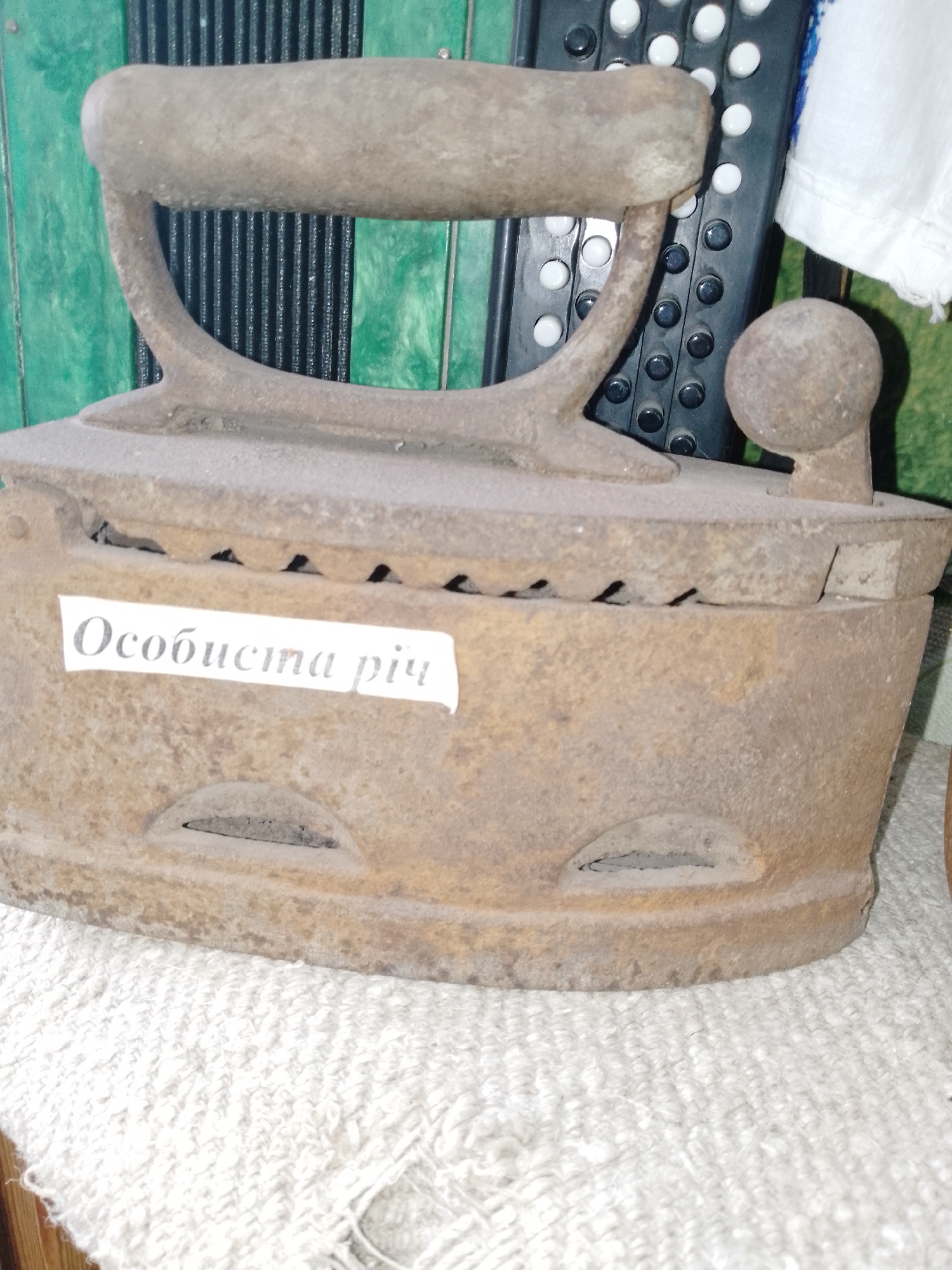 The image presents a close-up of an old, heavily rusted cast-iron charcoal or sad iron, displayed in an indoor setting in Ustyvytsia, Ukraine. The iron, the primary subject, exhibits significant wear, dust, and rust, indicating its age and disuse. It features a robust, curved handle and a hinged top cover with several small vents along its edges, designed for the release of smoke or heat from coals placed inside. The bottom portion of the iron also has two larger, semi-oval vents.
Affixed to the front side of the iron is a white paper label with text printed in black. The visible text reads: "Особиста річ". In Ukrainian, this translates to "Personal item" or "Personal belonging," suggesting the iron holds historical or sentimental value.
In the background, elements of the setting are partially visible. To the left, there's a bright green paneling or wall. To the right, behind the iron, parts of a black accordion or bayan with white and black keys can be seen. Further to the right, a piece of white, textured fabric, possibly a towel or traditional linen, is draped.
The scene depicts a static arrangement of objects, likely part of a historical or cultural exhibit, given the condition and context of the items. No activity is taking place, and the time of day or weather is not discernible. The composition emphasizes the historical artifact, evoking a sense of past domestic life and Ukrainian heritage.