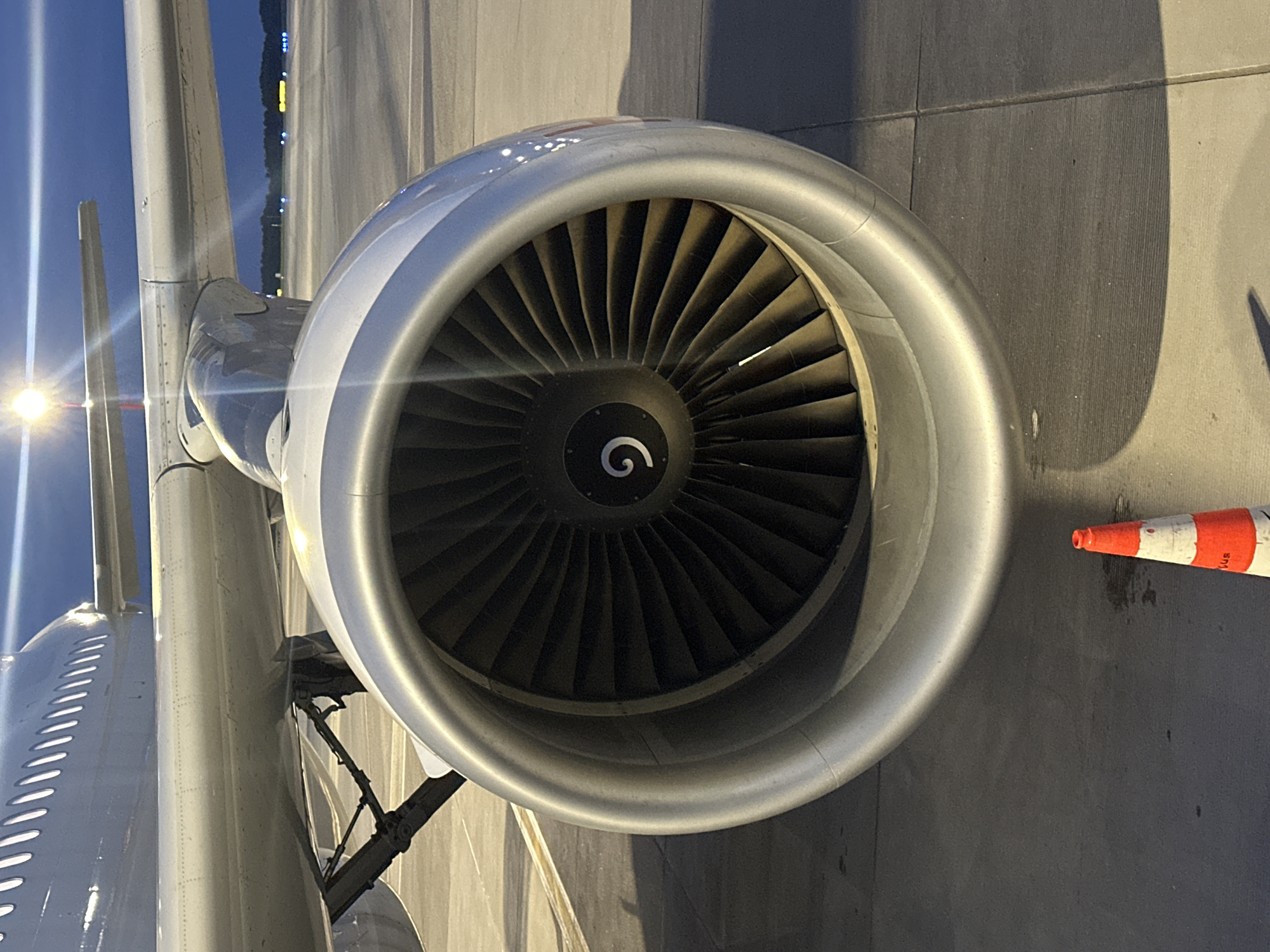 The image is a close-up shot of an airplane engine on an airport tarmac. The engine is silver and has visible fan blades inside. A portion of the airplane's wing and fuselage is visible above the engine. The tarmac is gray concrete, and a small orange and white traffic cone is visible in the lower right corner. In the background, distant lights suggest an airport environment at dusk or night. The scene is devoid of people. The image does not provide any specific clues or context to indicate its location in the city of Vilnius, Lithuania.