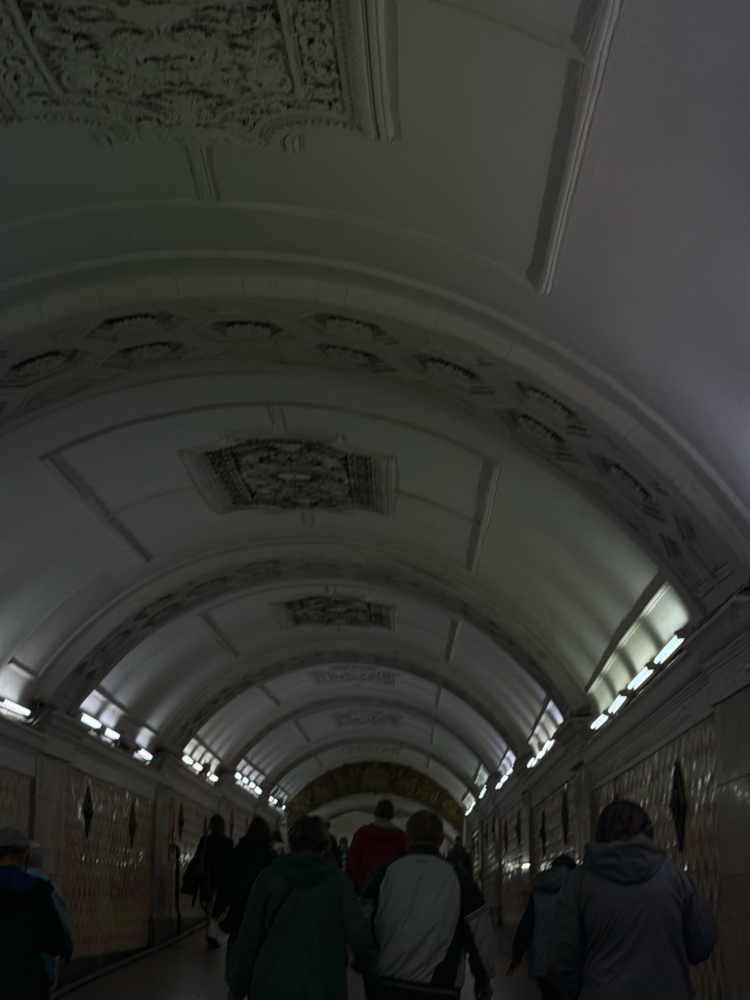 The image shows a dimly lit, arched pedestrian tunnel, likely a metro station underpass in Moscow, Russia. The ceiling of the tunnel features decorative plasterwork and inset lighting panels. Several people are walking away from the camera in the same direction, their figures silhouetted against the background. One person in the foreground on the right wears a dark gray hooded jacket and walks with their back to the camera. The walls of the tunnel are tiled with a patterned design.