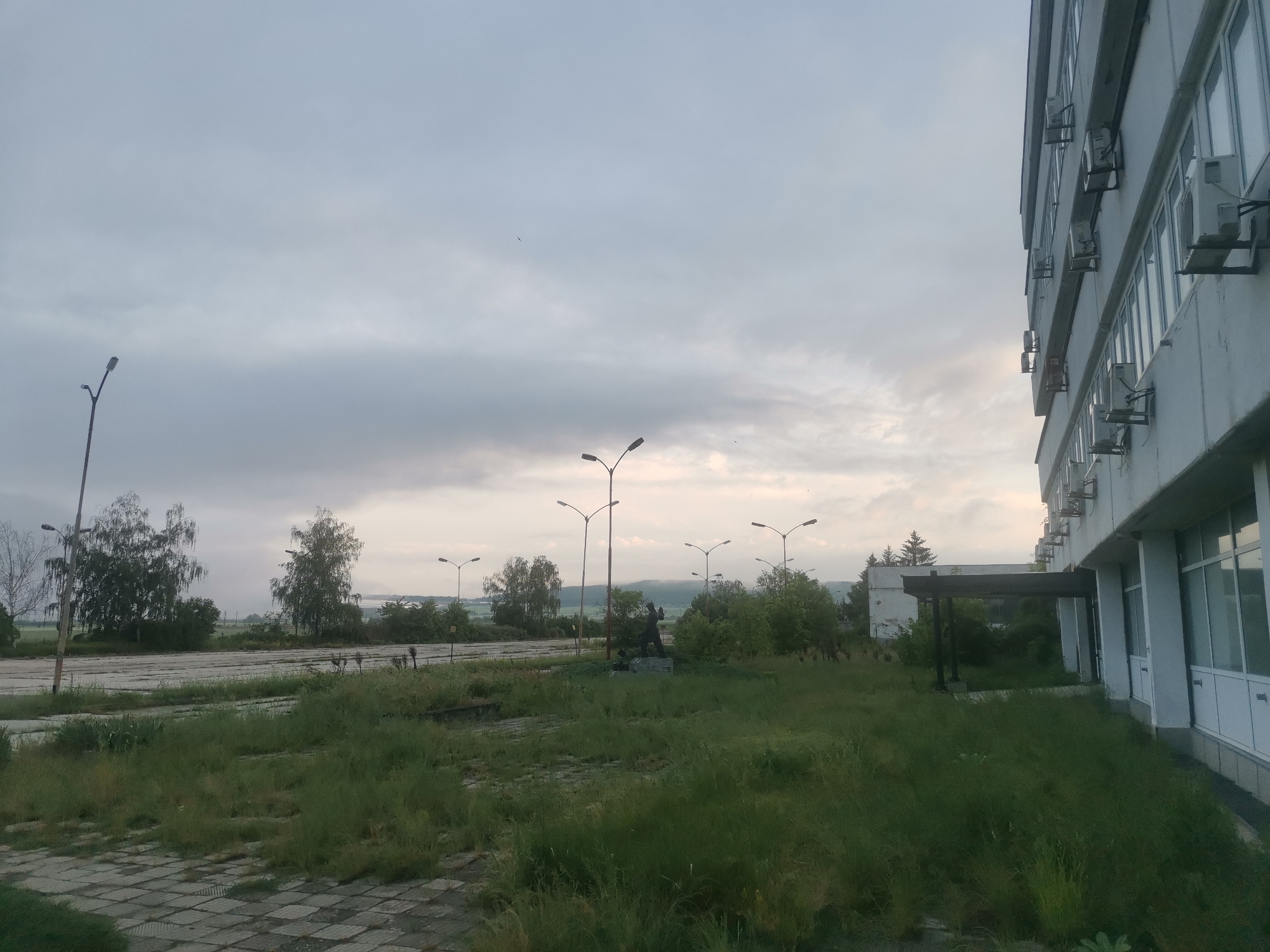 The image presents an overcast outdoor scene in Zhivkovo, Bulgaria, characterized by an aging building on the right and an expanse of overgrown land. The multi-story building, painted in light grey or white, features numerous windows and external air conditioning units affixed to its facade, indicating an older structure with more recent installations.
In the immediate foreground, a patterned paved area is partially obscured by tall, unkempt grass, which dominates much of the ground leading into the middle distance. Beyond this, a wide, cracked surface, possibly old asphalt or concrete pavement, stretches horizontally across the frame. Scattered throughout the scene are several tall streetlights, some with single lamps, others with twin fixtures.
In the mid-ground, a dark-colored statue, depicting a human figure on a pedestal, rises from the overgrown grass, surrounded by bushes. Behind the statue, a dark, covered walkway or structure is partially visible, along with a smaller, distant white building nestled among trees. Various trees, including what appear to be birches and some conifers, punctuate the landscape towards the horizon.
The sky is heavily clouded, displaying shades of grey with lighter patches near the horizon, suggesting either early morning or late afternoon. The overall impression is one of quietude and neglect, with no discernible human activity. No visible text is present within the image.