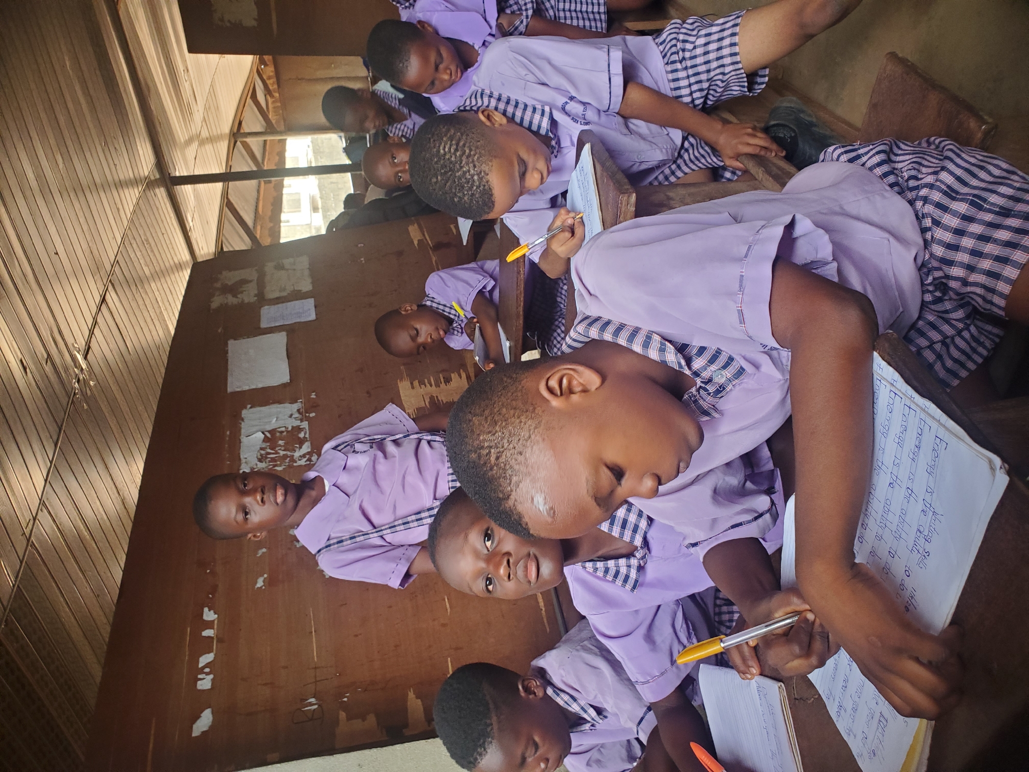 In a classroom located in Ukwu-Abwa, Nigeria, a group of elementary school children are seated at wooden desks, diligently writing in notebooks. Each child is dressed in a purple school uniform with a checked collar and plaid shorts. Their attention is focused on their writing tasks, as they use pens to inscribe text in their notebooks. The classroom is simply furnished with wooden walls and desks, reflecting a basic educational environment. The children are actively engaged in their schoolwork, illustrating a typical classroom setting in this Nigerian community.
