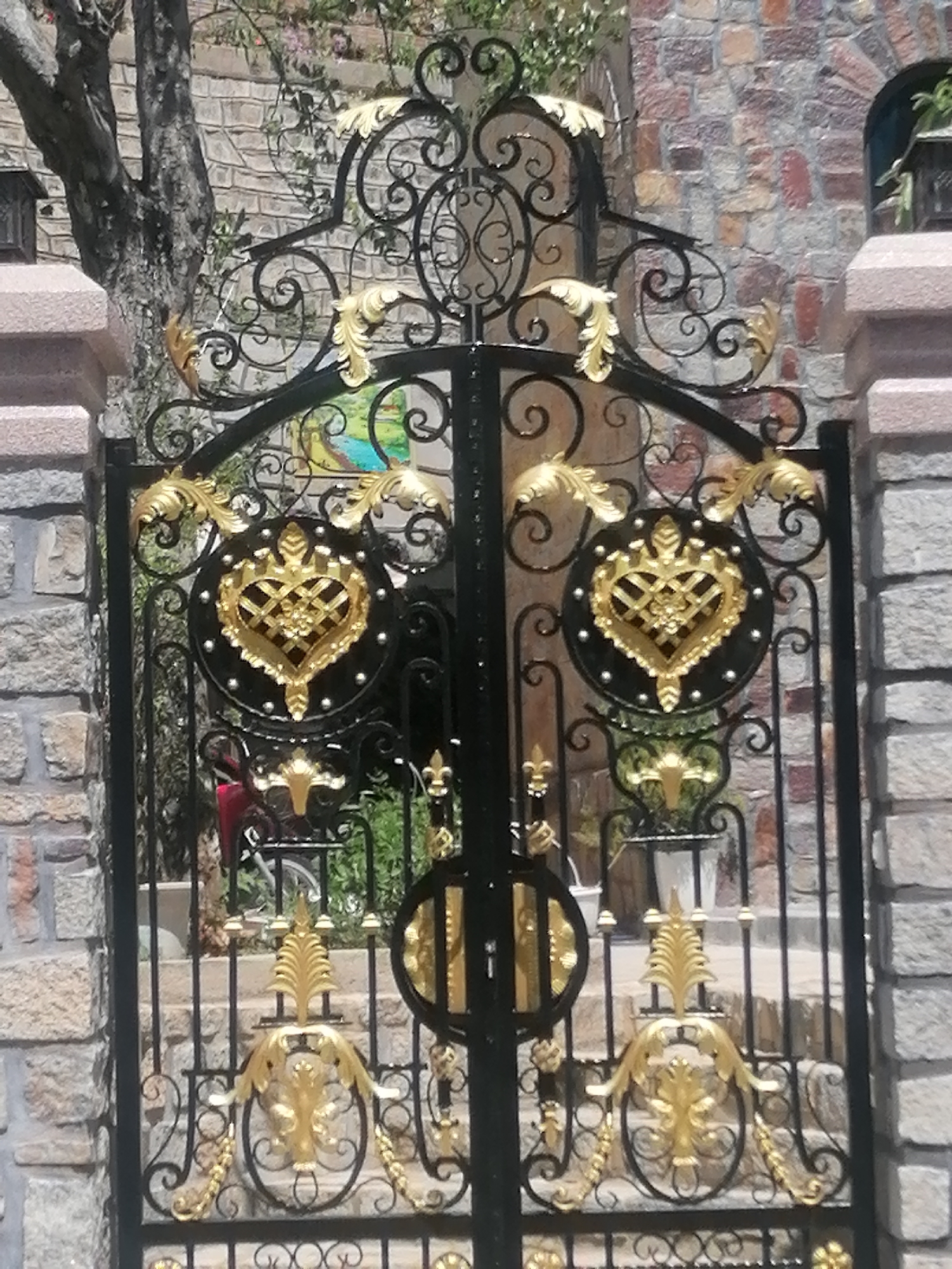 This is a photograph of an ornate black metal gate with gold accents, located in Da Lat, Vietnam. The gate features intricate scrollwork and decorative elements, including two large circular medallions with gold heart designs. The gate is set within stone pillars and a stone wall. Behind the gate, there is a staircase and lush greenery, with a hint of a tree on the left side of the frame. The style of the gate suggests it may be part of a residence or establishment with a grand or traditional aesthetic.