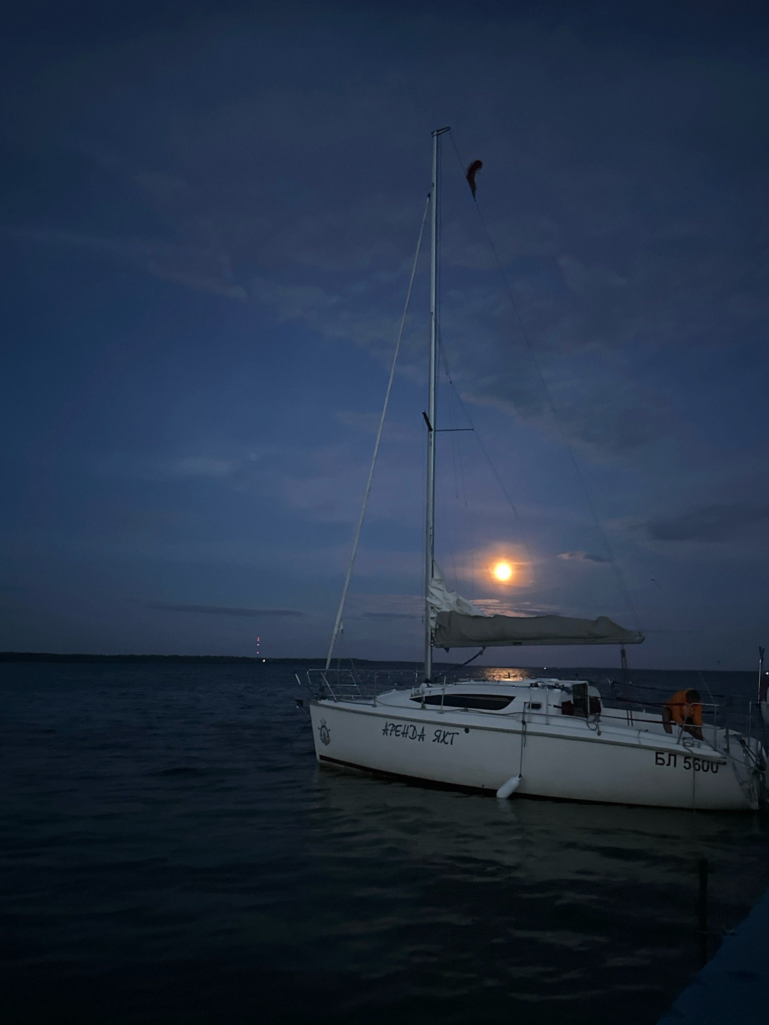 A white sailboat is docked on a body of water under a dark blue, cloudy sky with a full moon partially obscured by clouds. The sailboat has a tall mast with sails furled, and the text "АРЕНДА ЯХТ" and "БЛ 5600" are visible on its hull. An orange life jacket is visible on the deck of the boat. In the distance, a faint light is visible on the horizon, possibly a lighthouse or tower. The water is calm with gentle ripples. The scene appears to be set at dusk or night. The location is indicated as Нарацкі сельскі Савет, Belarus.