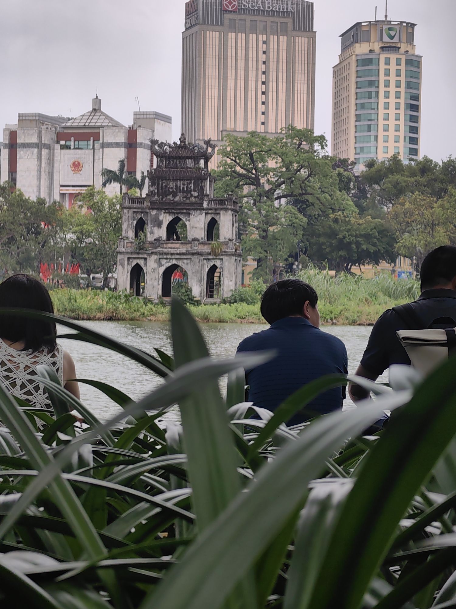 The image presents a serene view across Hoan Kiem Lake in Hà Nội, Vietnam, on what appears to be an overcast day.
In the center of the lake stands the historic Turtle Tower (Tháp Rùa), a weathered, multi-tiered stone structure featuring multiple arched entrances on its lower level and an ornate roof. Lush green foliage surrounds the tower and the distant banks of the lake.
The background showcases a blend of modern and traditional Vietnamese architecture. To the left, a large, light-colored building with a distinctive angular roof and a prominent red emblem (resembling the Vietnamese national emblem) is visible. Behind this, a tall skyscraper prominently displays "SeABank" and partially "BRG Group" near its top. Further to the right, another modern high-rise building, lighter in color with numerous windows and a green logo at its peak, adds to the urban skyline.
In the immediate foreground, out-of-focus green plants create a natural frame for the scene. Through these plants, three individuals are seen from behind, appearing to be observing the view across the lake. To the left, a woman with short dark hair wears a white, intricately patterned top. To her right, two men are seated, one in a dark blue polo shirt and the other in a dark top with a light-colored backpack. Their posture suggests a moment of leisure, enjoying the iconic landmark and the surrounding cityscape. The soft, diffuse lighting indicates a cloudy sky, contributing to a calm and reflective atmosphere.