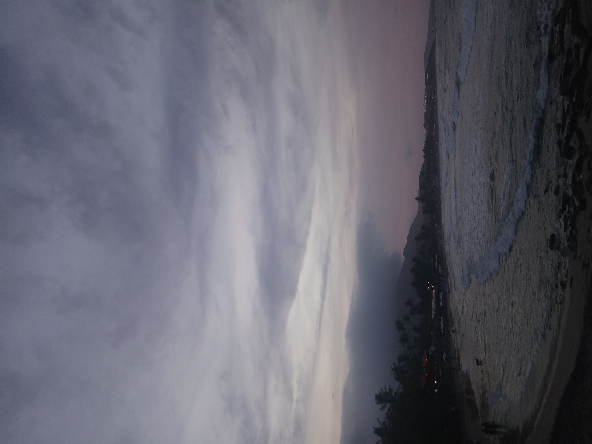 The image shows a dusk or dawn view of a beach in Juan Griego, Venezuela. To the left is a sky filled with striated clouds. To the right, the ocean waves break on the shore, with hints of city lights and palm trees lining the beach. There are silhouettes of people in the lower portion of the image.