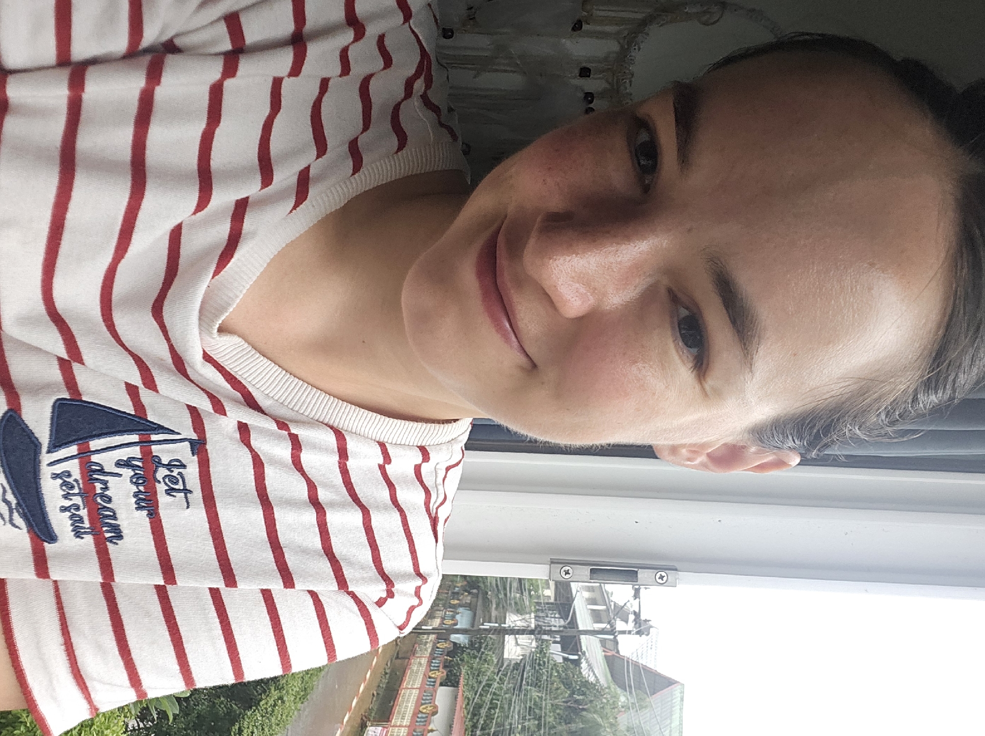 A young woman with fair skin, freckles, and dark hair pulled back is taking a selfie in Kathu, Thailand. She is wearing a white t-shirt with horizontal red stripes and an embroidered sailboat with the text "Let your dream set sail." She is positioned in front of a window, with a glimpse of a residential street and lush green trees visible outside. The window frame is white and has a metal latch. The overall lighting suggests it is daytime, possibly overcast.