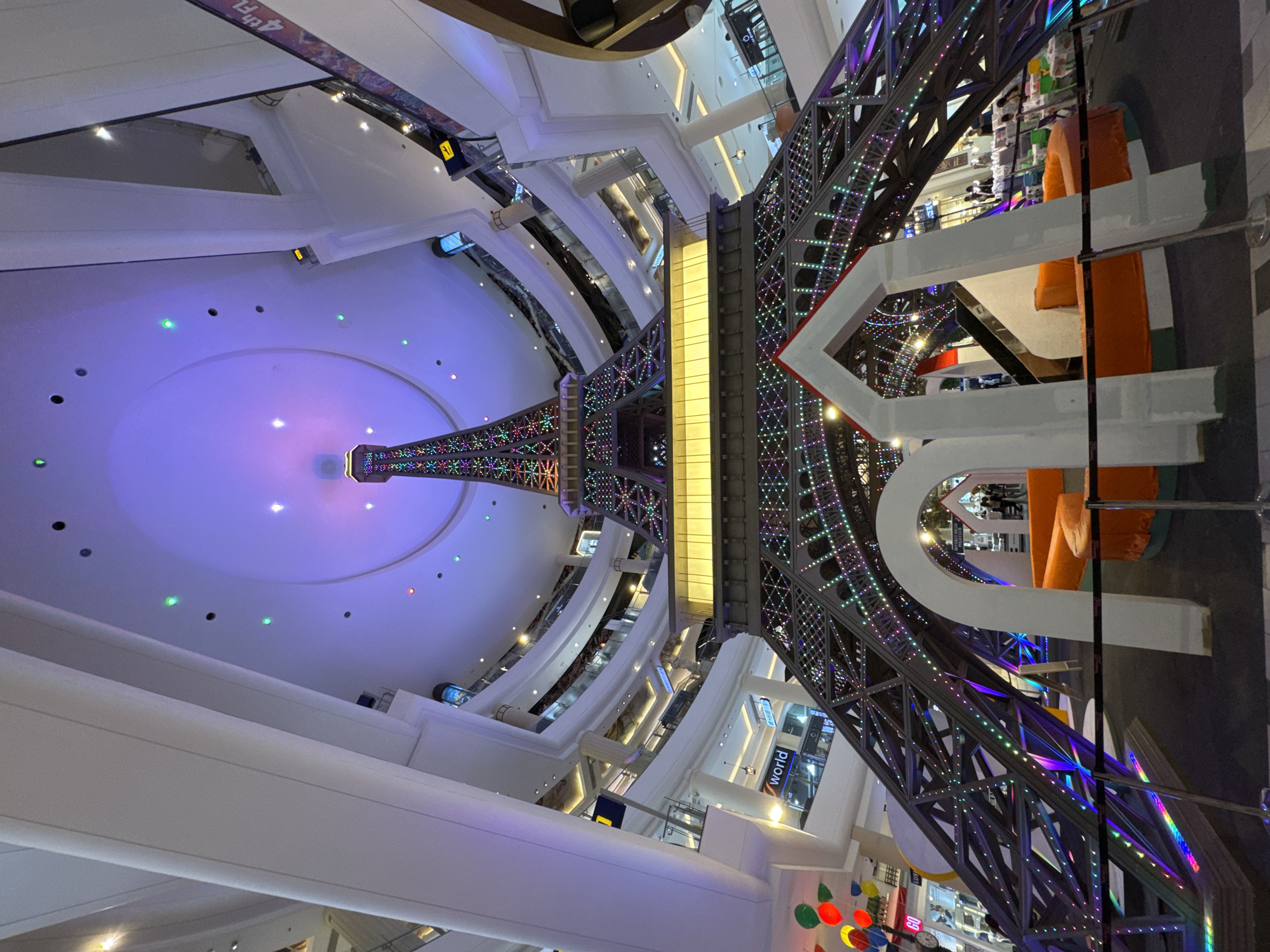 An indoor, multi-level shopping mall in Pattaya City, Thailand, features a large, illuminated replica of the Eiffel Tower as a central decorative element. The tower, adorned with colorful LED lights, is situated in a spacious atrium with multiple floors visible in the background, each with escalators and railings. The ceiling of the mall is a large, domed structure with purple and blue lighting and scattered green and pink spotlights. In the foreground, part of the base of the Eiffel Tower replica is visible, with a decorative archway structure made of white material and orange seating. Several people are present in the background, shopping or walking through the mall. Balloons in red, yellow, and green are clustered on the left side of the image. The overall scene depicts a festive and brightly lit commercial environment.