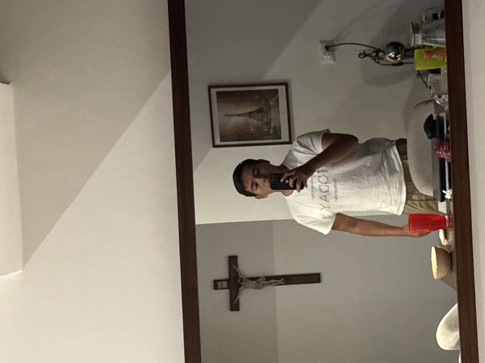 The image shows a mirror's reflection in San Bernardino Tlaxcalancingo, Mexico. A person is taking a photo of themselves with a phone, wearing a white t-shirt and khaki shorts, holding a red plastic cup. In the background is a wall with a framed picture of the Eiffel Tower, an electrical outlet, a crucifix, and a shelf holding various items. The environment appears to be an interior space with white walls and a light-colored ceiling. A brown wooden frame surrounds the mirror.