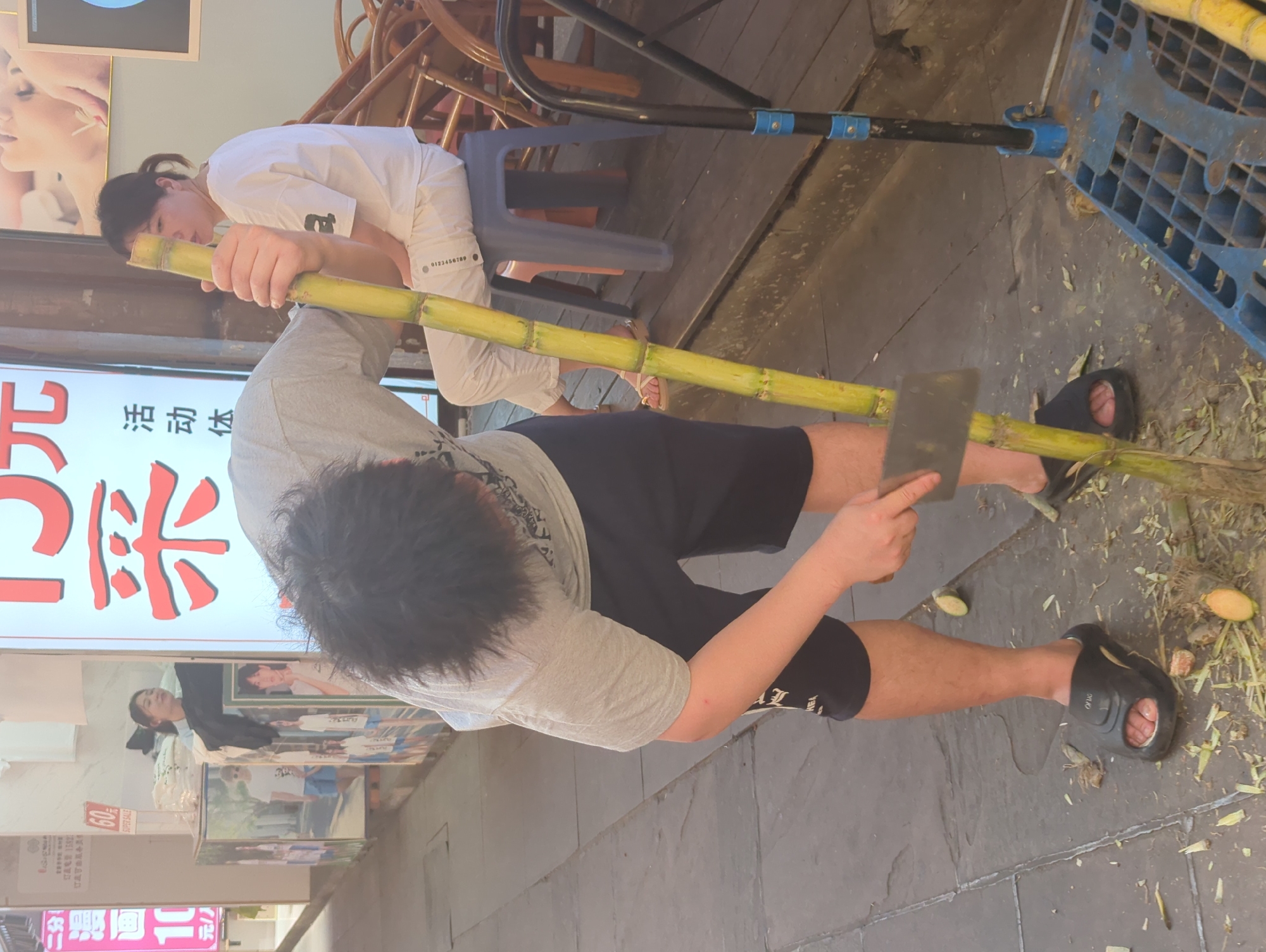 The image captures a vibrant street scene, likely in an urban area of Yuzhong District, China, centered around the preparation of sugarcane. In the foreground, a person with dark hair, wearing a grey t-shirt and black shorts with black sandals, is actively peeling a long sugarcane stalk. They hold a large, flat-bladed knife or cleaver in their right hand, using it to strip away the outer layer of the cane. Sugarcane husks and some fruit peels are scattered on the grey tiled ground around their feet.
In the mid-ground, another person, appearing to be female, is seated on a dark blue plastic stool. She wears a white t-shirt and white pants with light-colored sandals. Her head is bowed slightly as if watching the activity or resting. Behind her, part of a wooden chair or table structure is visible.
The setting appears to be an outdoor or semi-enclosed commercial space, possibly a storefront or food stall. Brightly lit signs with Chinese characters are prominent on the left, one displaying a large red character that looks like part of "采" (cǎi). Another sign advertises "60 SUPER SALE." Posters and advertisements depicting people are also visible on the wall behind the seated individual. A numerical sequence "0123456789" is printed on the white pants of the seated person.
Other visible text from the signs includes fragments like "二分", "订正可能服务员", "漫画 1", "元/人", "活动体", and "HEA". The scene is well-lit, suggesting either daytime or a brightly illuminated evening environment.