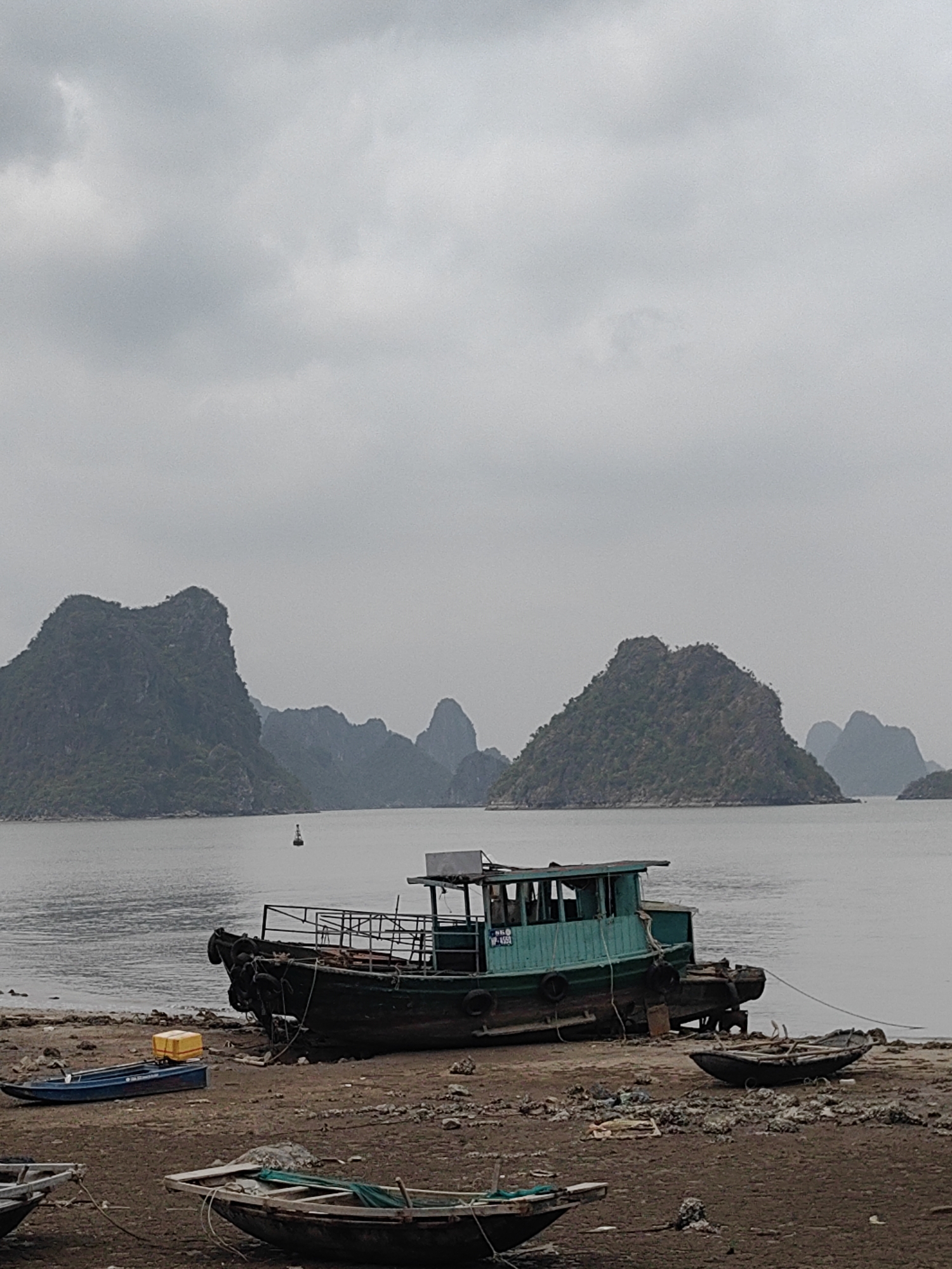 The photo depicts a seascape scene in Hải Phòng, Vietnam. The scene is overcast with a grey sky. The sea stretches to the horizon, with several karst islands rising from the water. In the foreground, boats rest on the shore. One is a larger, green-painted vessel. Smaller boats are scattered around. Debris litters the beach.