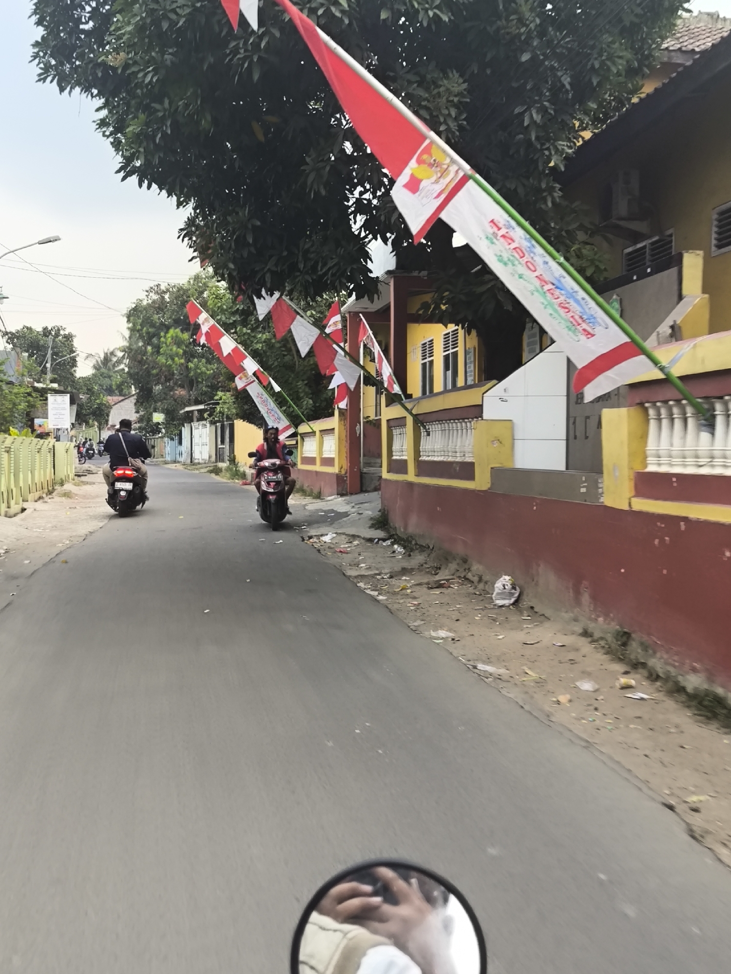 A low-angle, forward-facing view from a moving motorcycle on a paved street in Cilegon, Indonesia. The street is lined with buildings and vegetation. The dominant colors are gray asphalt, yellow and red buildings, and green foliage. Indonesian flags, red and white, are prominently displayed along the right side of the street, attached to poles. Some flags have text and emblems. Two motorcycles are visible on the road ahead, each with a rider. The motorcycle on the left is black and ridden by a person wearing dark clothing. The motorcycle on the right is dark red and ridden by a person wearing a red shirt. In the foreground, the reflective mirror of the motorcycle on which the photo is taken shows a partial reflection of the rider's hand. The environment appears to be a residential or commercial area, with some litter visible on the side of the road. The sky is overcast.