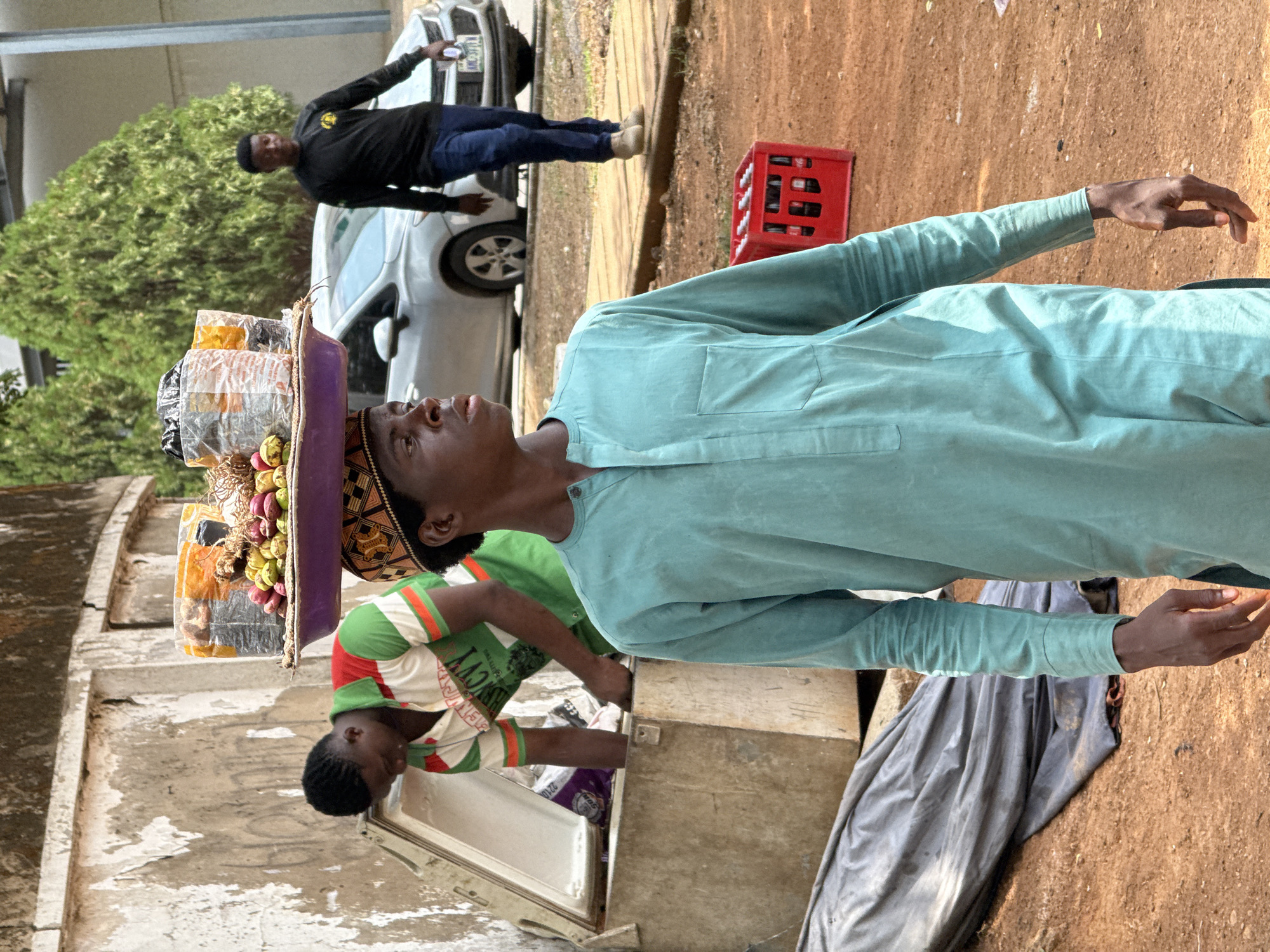 A man in a light blue robe and a patterned cap stands with a tray of goods balanced on his head. He is looking up and has his arms outstretched. Next to him, a younger man wearing a green, white, and red striped shirt is reaching into a weathered chest refrigerator. Behind them, a third man in a black shirt and jeans stands near a silver car. A red crate sits on the ground nearby. The scene appears to be a street in Abuja, Nigeria, evident from the visible architecture, ground texture, and general context.