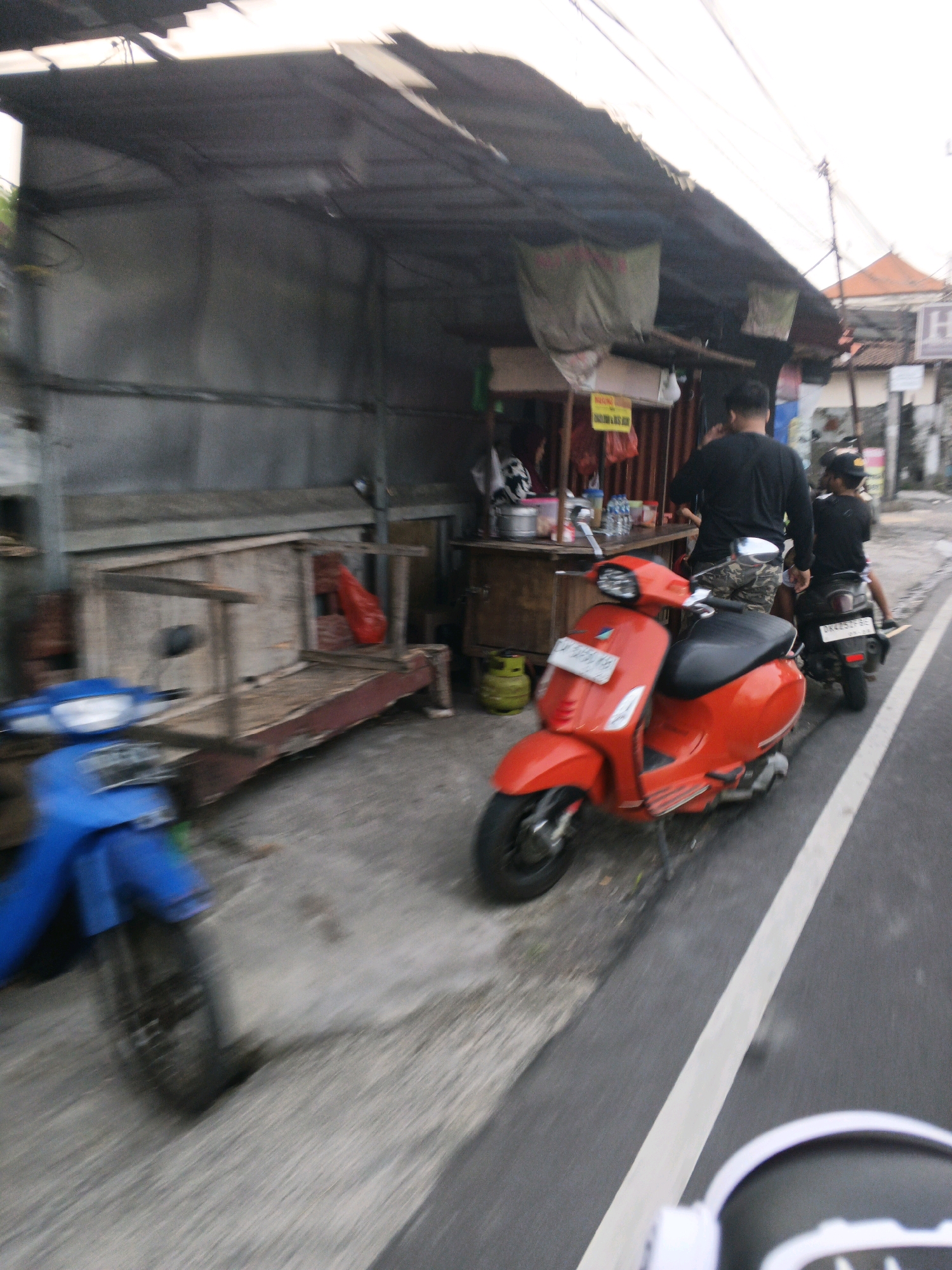 The image depicts a street scene in Denpasar, Indonesia, characterized by a roadside stall and parked motorbikes. A red Vespa scooter is prominently displayed in the foreground, parked on the asphalt road. Behind it, a man wearing a black shirt and patterned shorts stands near the stall, while another person, also in a black shirt and a cap, sits on a dark-colored motorbike. The stall itself appears to be a simple structure with a corrugated metal roof, offering various items that are not clearly identifiable due to the image's blurriness. To the left of the stall, a blue motorbike is partially visible. The environment suggests a typical urban setting in Denpasar, with a sidewalk area, utility poles and wires overhead, and buildings in the background. The overall impression is one of a bustling street with local commerce and transportation.