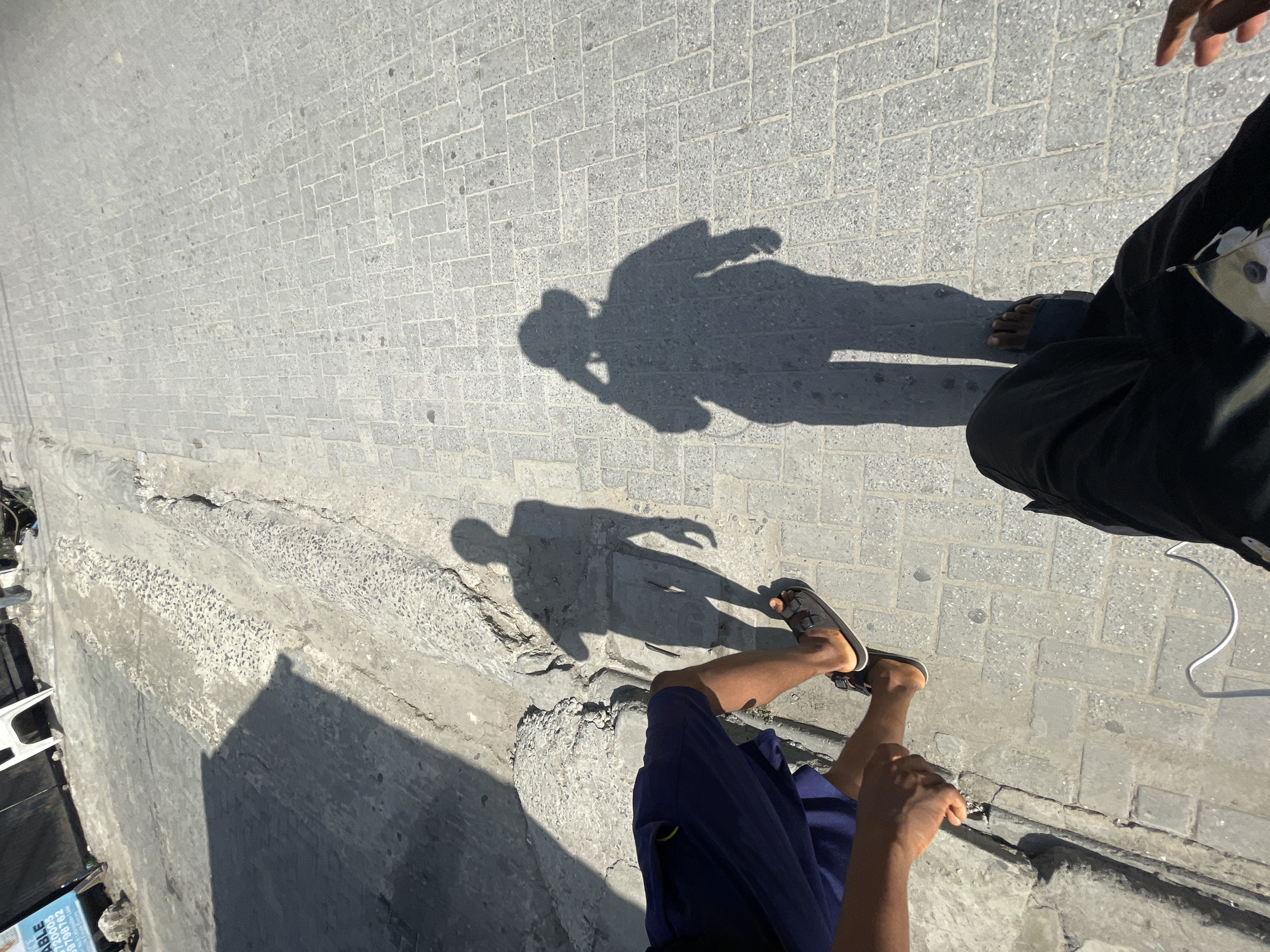 The image presents an overhead, high-angle view of two individuals on a light gray paved surface, likely a street or walkway, under bright sunlight. The strong illumination creates distinct, sharp shadows.
In the foreground, towards the bottom right, are the legs of one person wearing blue shorts and dark sandals. Their hand is also visible near the bottom edge. This person casts a prominent shadow on the textured, interlocking brick pavement. The shadow clearly outlines a human figure, possibly with one arm raised or holding something near their head.
Further back and to the right is another person, primarily visible from the waist down, wearing dark trousers or a long garment. Their feet appear to be bare or in minimal dark footwear, resting directly on the pavement. A small white object, resembling a mask or small pouch, is attached to their dark clothing. This individual also casts a shadow onto the ground, visible towards the upper left. A white cable runs from the right side, extending near their feet.
To the left of the scene, a rugged concrete ledge or curb runs across the frame, showing a coarse, exposed aggregate texture. Below this ledge, there's a lower area containing debris, including what appears to be a white plastic chair and other detritus. On a blue and white object among the debris, partially visible text reads "ABLE," "3000," and "0970182."
The sharp shadows indicate a clear, sunny day, likely around mid-day. The overall scene suggests an outdoor, urban or semi-urban setting in Iranla, Nigeria.
