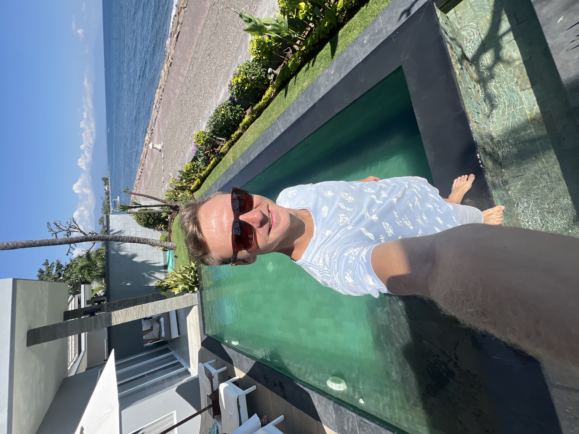A man takes a selfie by a pool overlooking the ocean. He is wearing sunglasses and a light blue t-shirt with a white pattern. The pool is a bright green color and is bordered by dark gray tiles. Behind the pool, there is a lawn with lush green plants and a stone wall. Beyond the wall, a beach stretches to the ocean, with mountains visible in the distance under a clear blue sky. The location is identified as Buleleng, Indonesia.