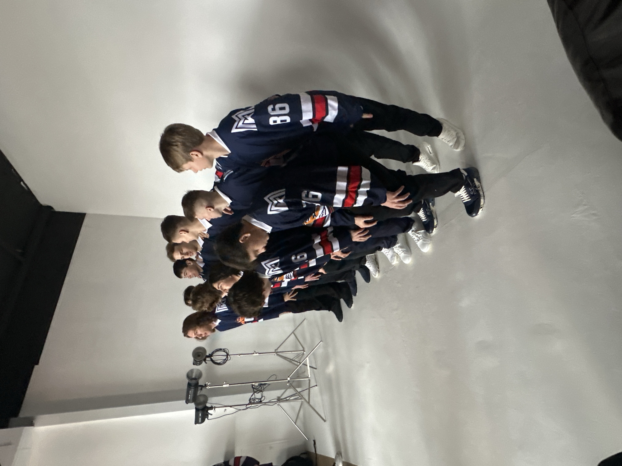 A group of approximately eight young people wearing similar navy blue hockey jerseys and dark pants are standing in a line in what appears to be a photography studio. The jerseys have white and red stripes across the sleeves and a logo on the front. The person at the front of the line has the number "86" on their back. The line of individuals is standing on a white floor and facing towards the right. A light stand with multiple lights is visible in the lower left corner of the frame. The location is Unknown, Unknown.