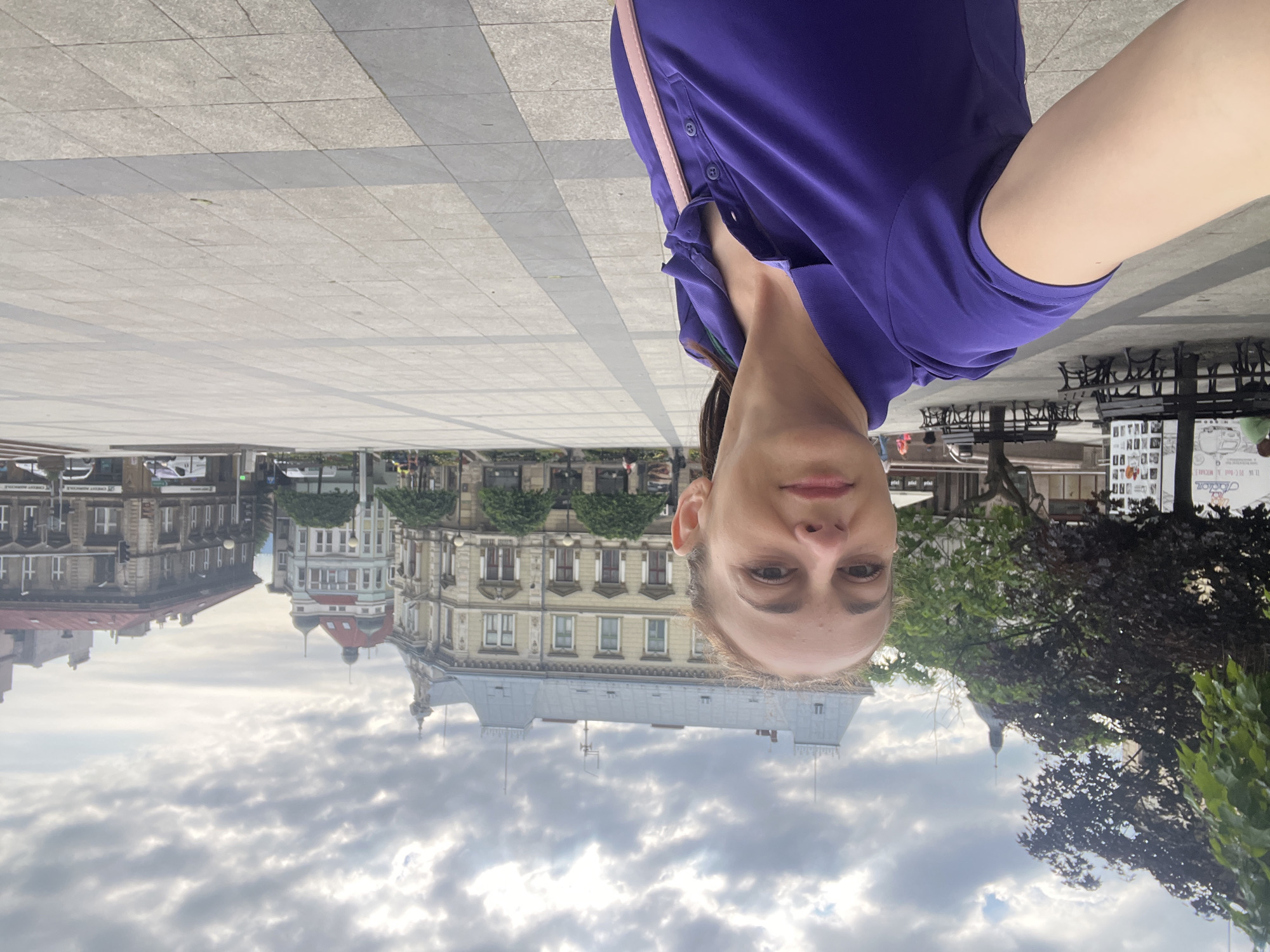 A woman in a purple polo shirt takes a selfie in a city square. She is in the foreground, looking towards the camera with a neutral expression. Her right arm is extended, holding the phone. Behind her, the square is paved with light-colored tiles and gray borders. Several trees are planted in rows. In the background, there are multi-story buildings with light-colored facades, some with red roofs, and architectural details such as windows and decorative elements. The sky is overcast with clouds. Benches are visible along the edge of the square, and there are some vehicles and a few people in the distance. The scene suggests an urban public space, likely a plaza or main square.